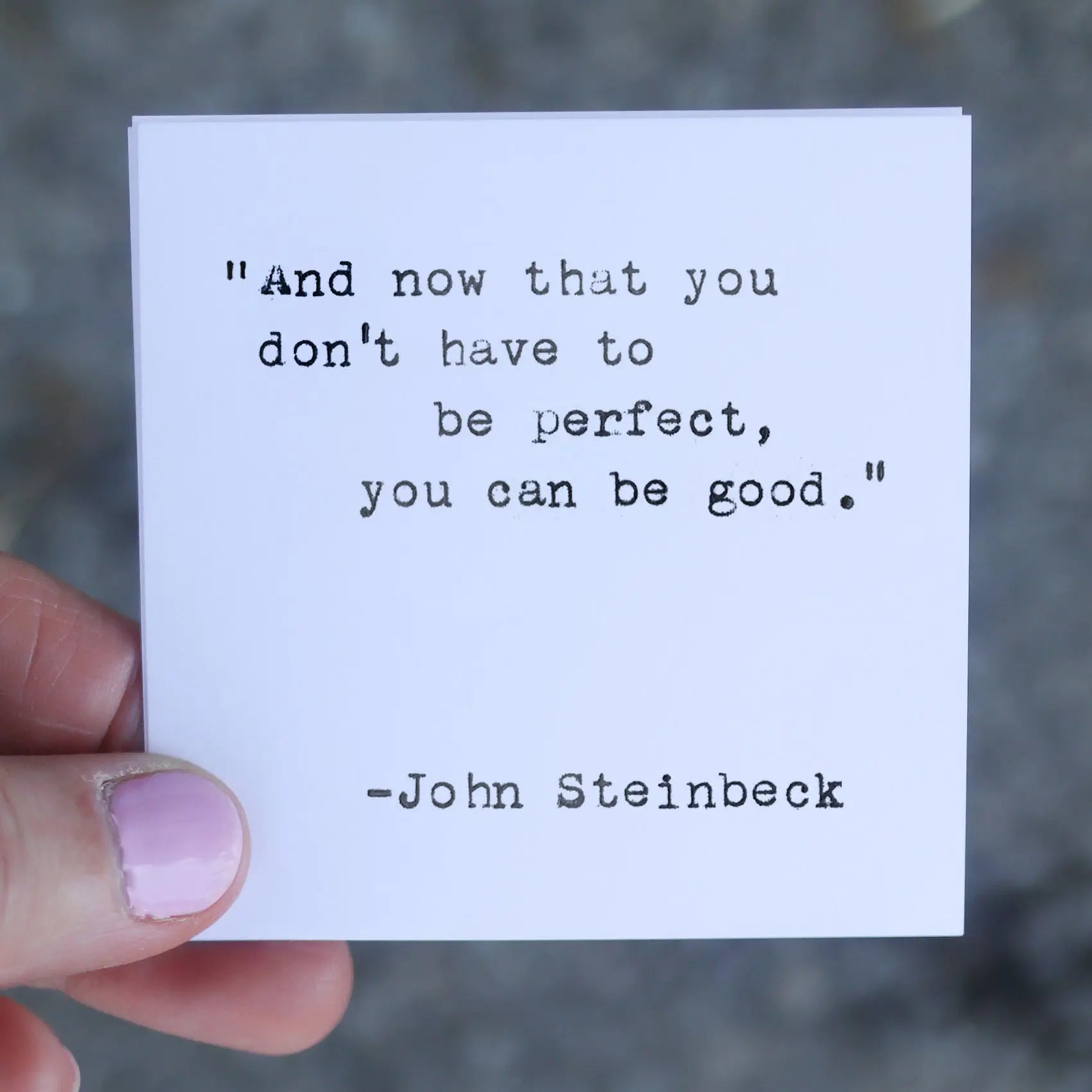 A finger holds a card with a John Steinbeck quote printed in typewriter font.