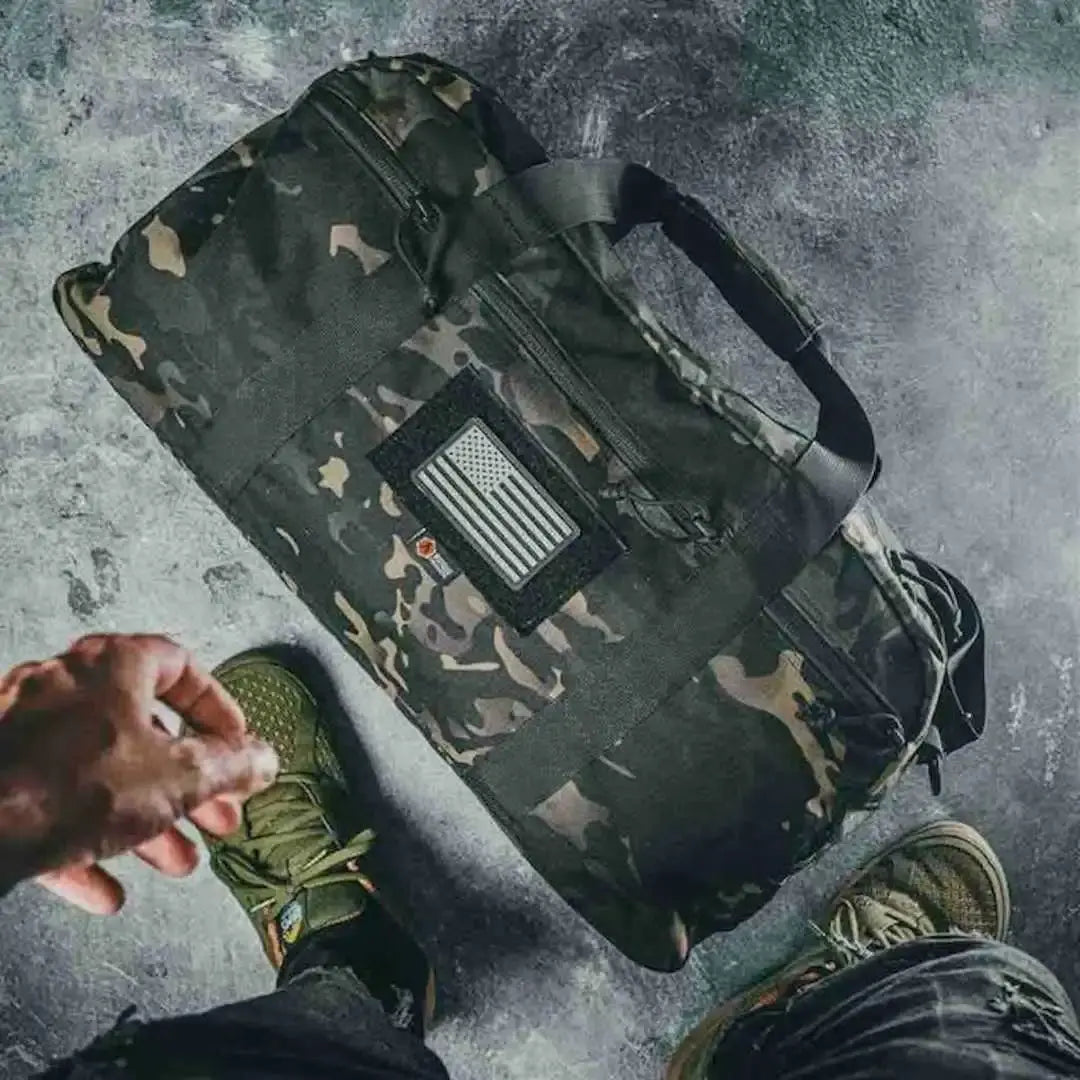 Camouflage duffle bag with an American flag patch on a textured surface.