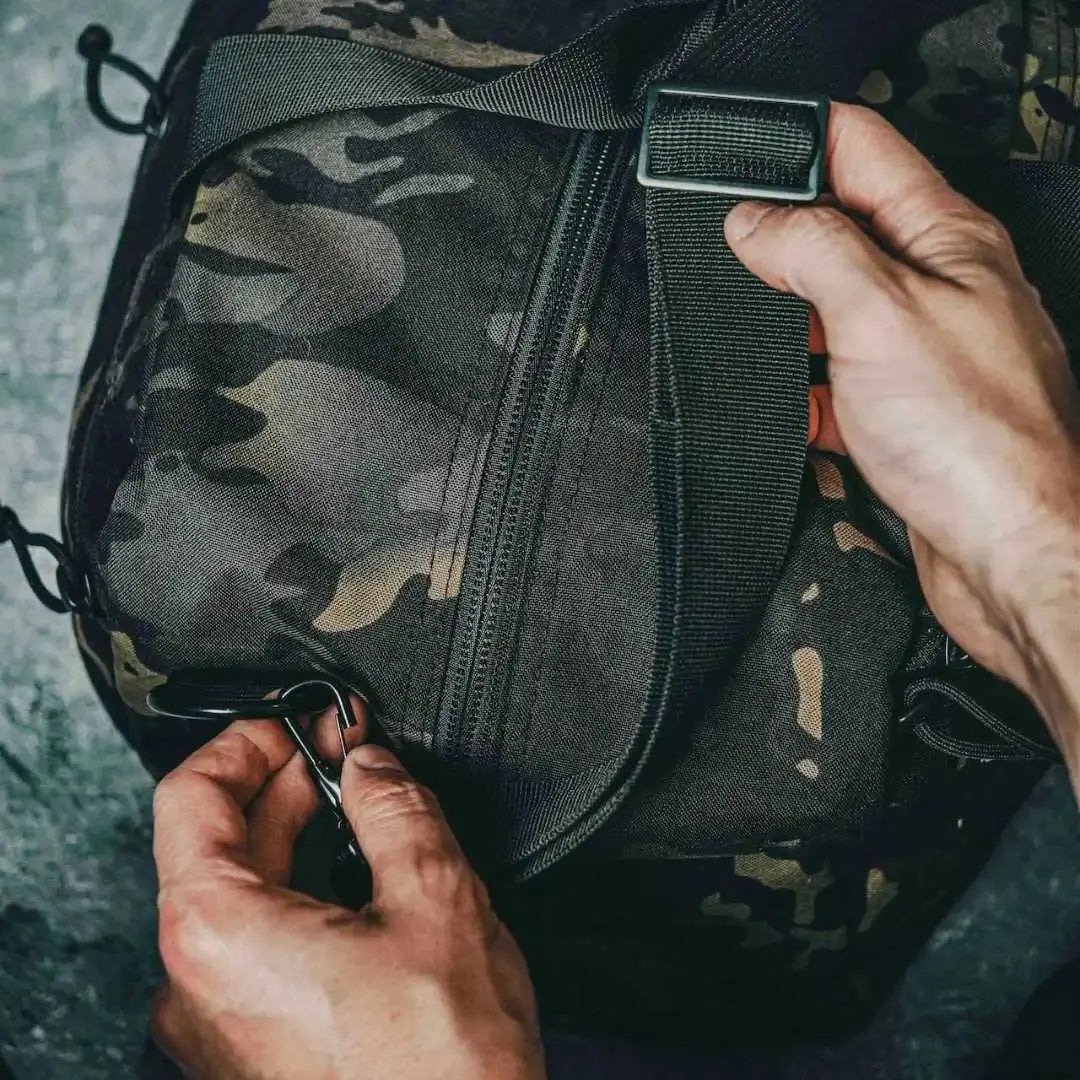 Hands adjusting a black camouflage 72-hour duffle bag with zipper and strap.