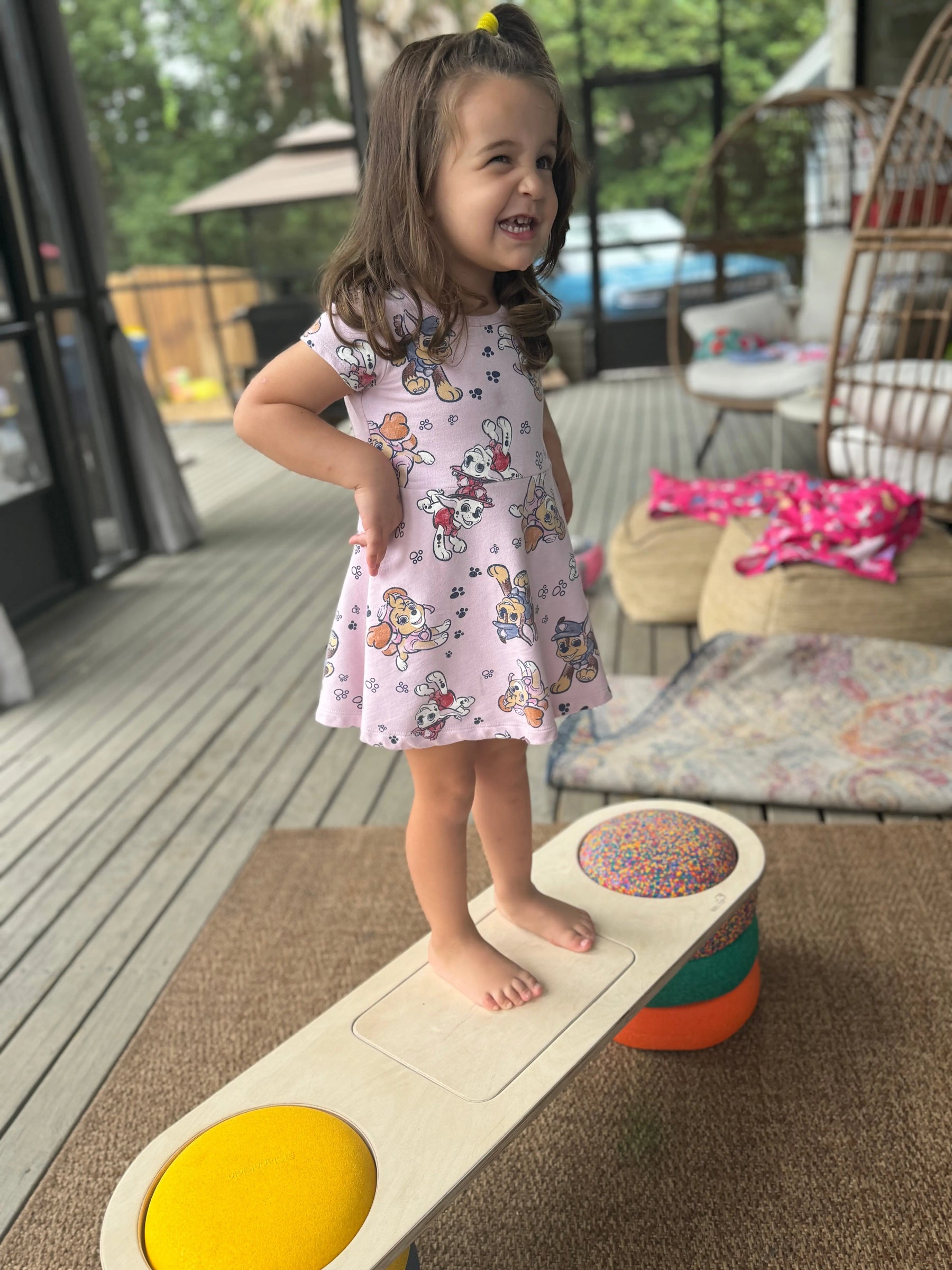 Young child in a Paw Patrol dress standing on a wooden balance board with colorful round inserts.