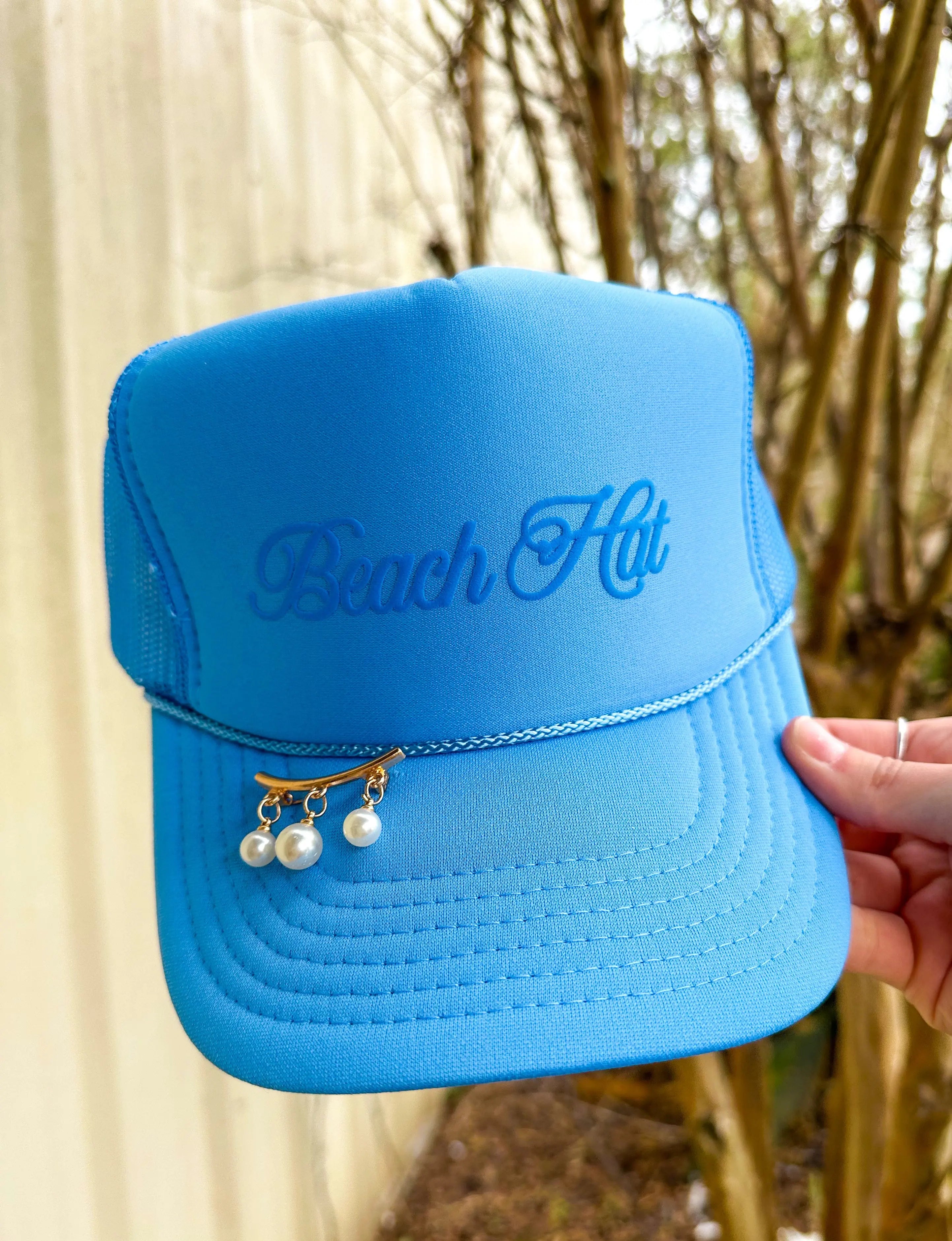 Columbia blue trucker hat with "Beach Hut" cursive text and pearl dangle accessory on brim held by a hand.