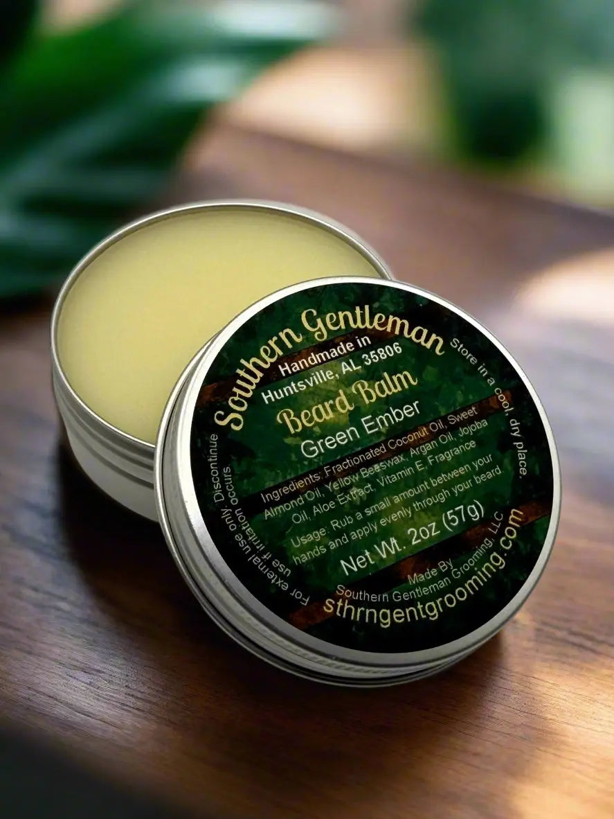 Open tin of Southern Gentleman Beard Balm 'Green Ember' on a wood surface with blurred green plant background.