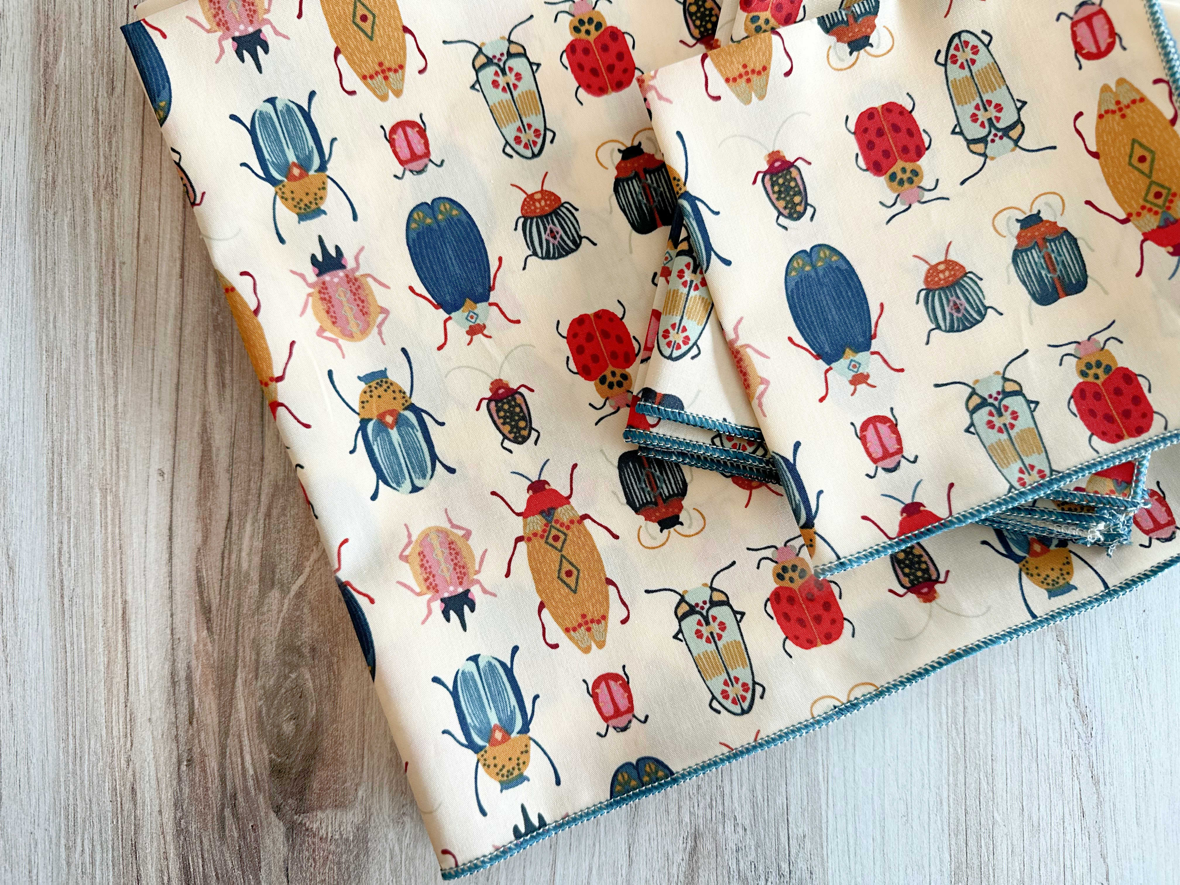 Cream cotton napkins with colorful beetle print and blue zig-zag trim, set of four.