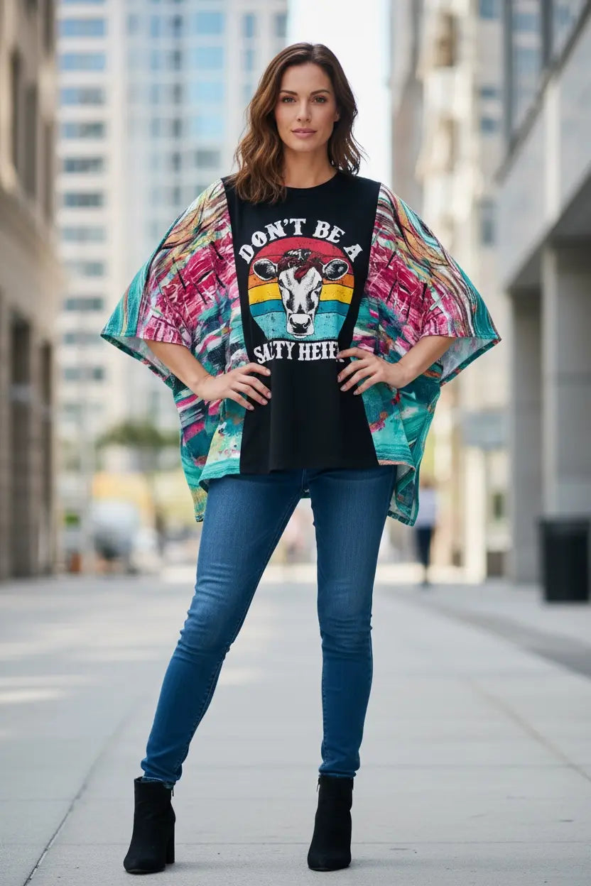 Woman in a boho poncho tee with a cow graphic, blue jeans, and boots standing outdoors.