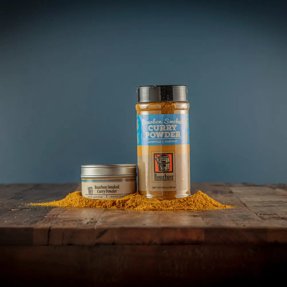 Two containers of Bourbon Smoked Curry Powder on a wooden surface with spilled powder.