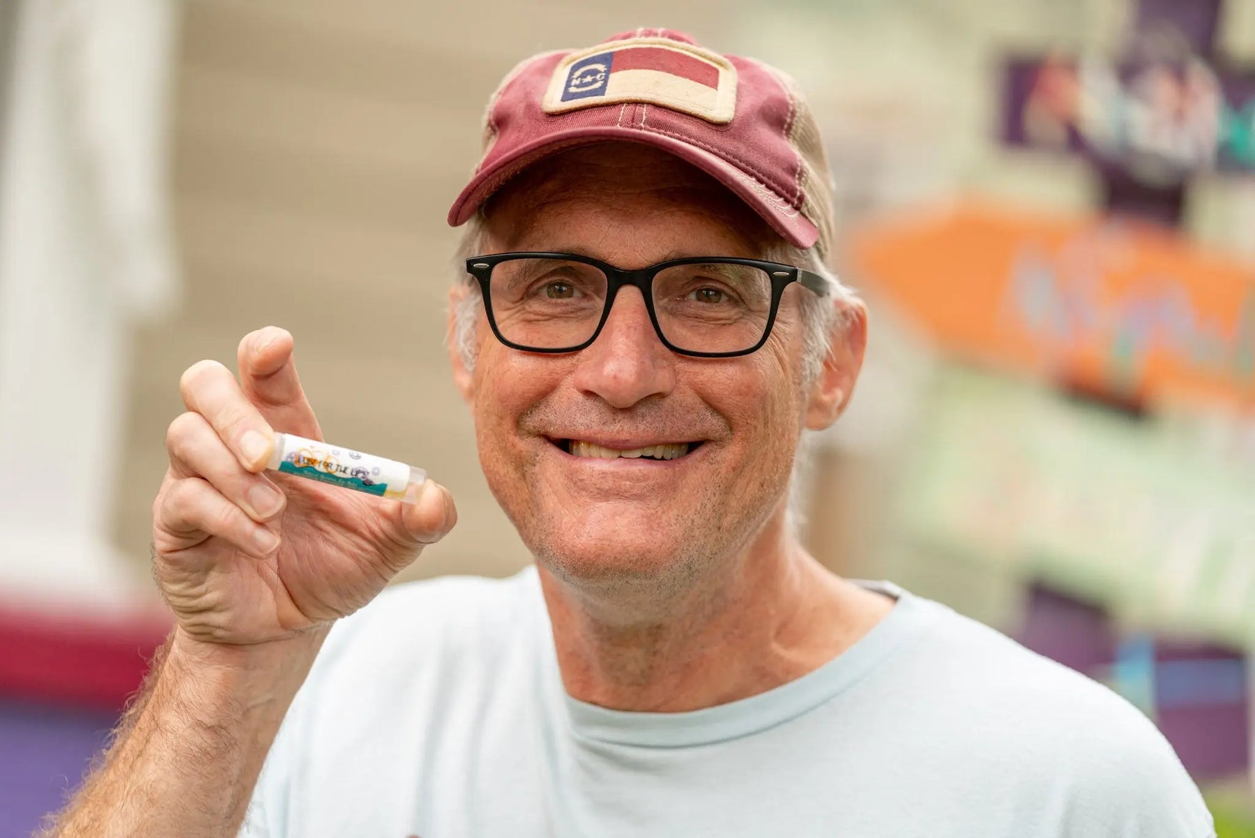 Man with glasses and hat holding "COASTING COCONUT SPF 15 LUBE FOR THE LIPS" lip balm.