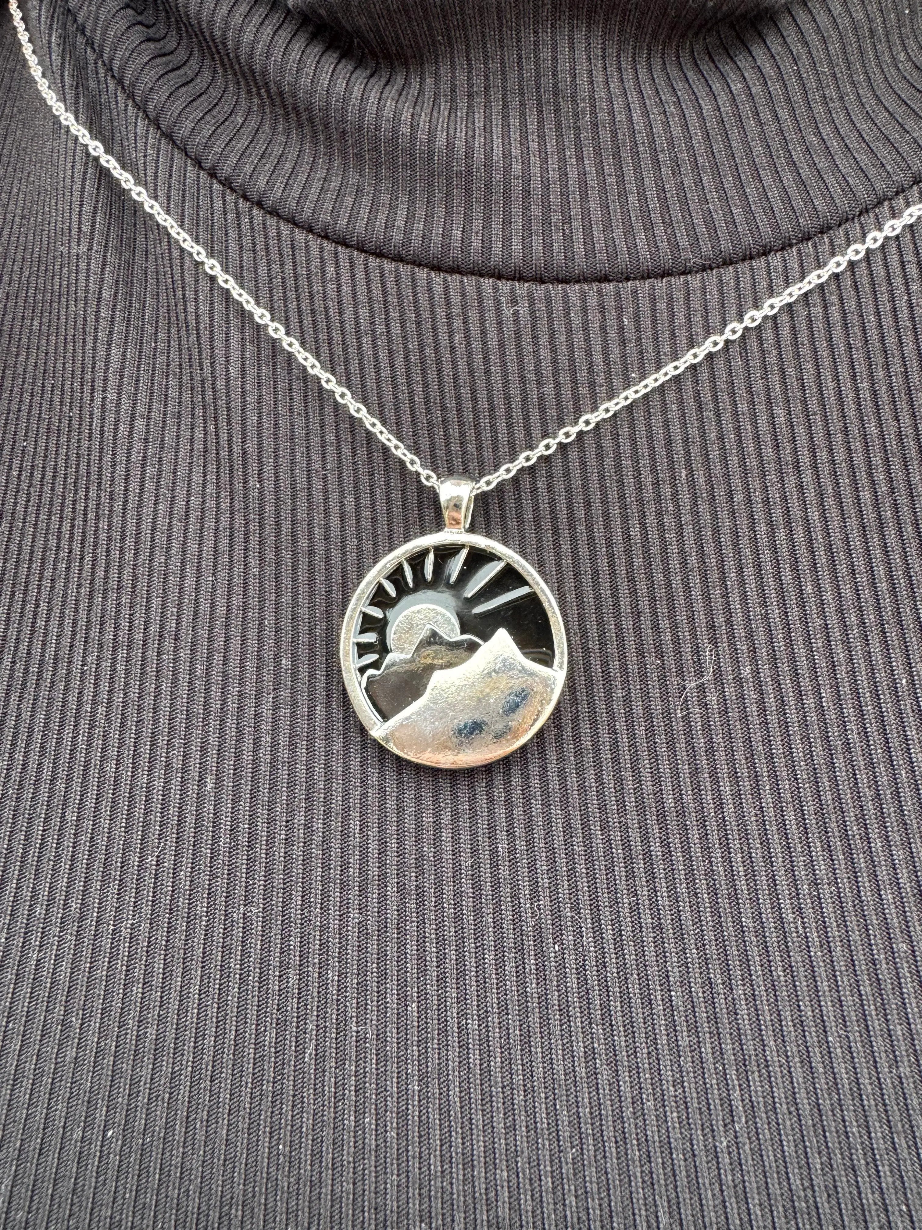 Silver mountain necklace: round pendant with mountain range, sun and navy background on a fine chain against black turtleneck.