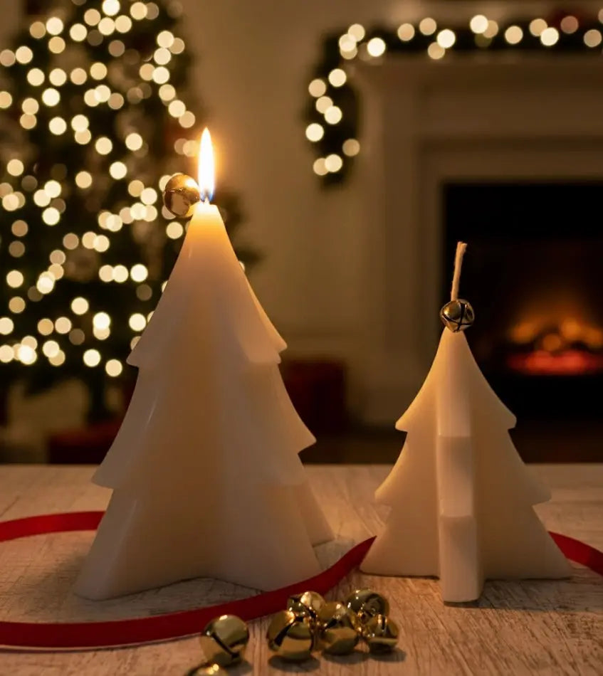 Two white Christmas tree candles, one lit, with bells and red ribbon on a wooden table.