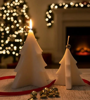Two white Christmas tree candles, one lit, with bells and red ribbon on a wooden table.