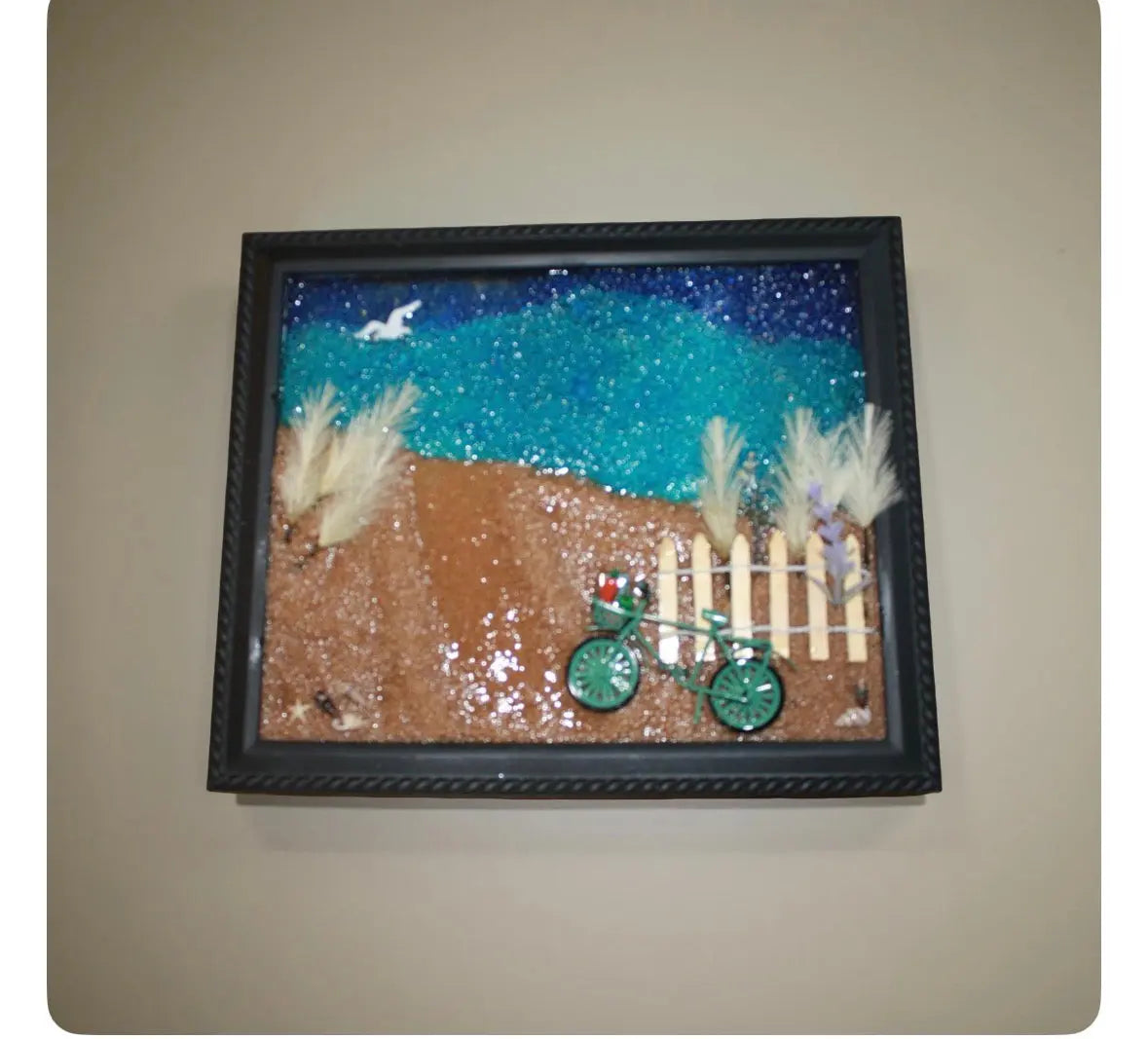 Framed resin glass artwork: coastal scene with bicycle, fence, and beach grass. Blue ocean, brown sand, green bicycle, and white fence.