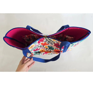 Open colorful floral tote bag with blue straps held by a hand.