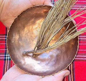 Handcrafted copper bowl in hands with pine needles on red plaid. Artisan rustic home decor.