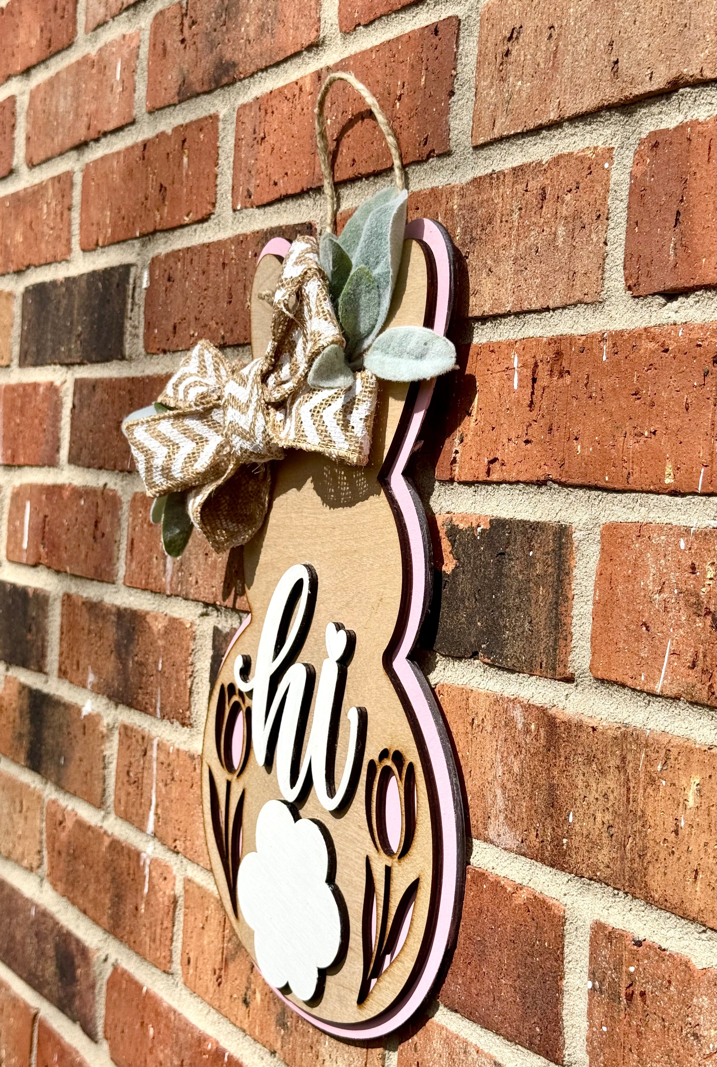 Bunny door hanger with bow on a brick wall. Wooden spring decor with "hi" message, tulips, and pink trim.