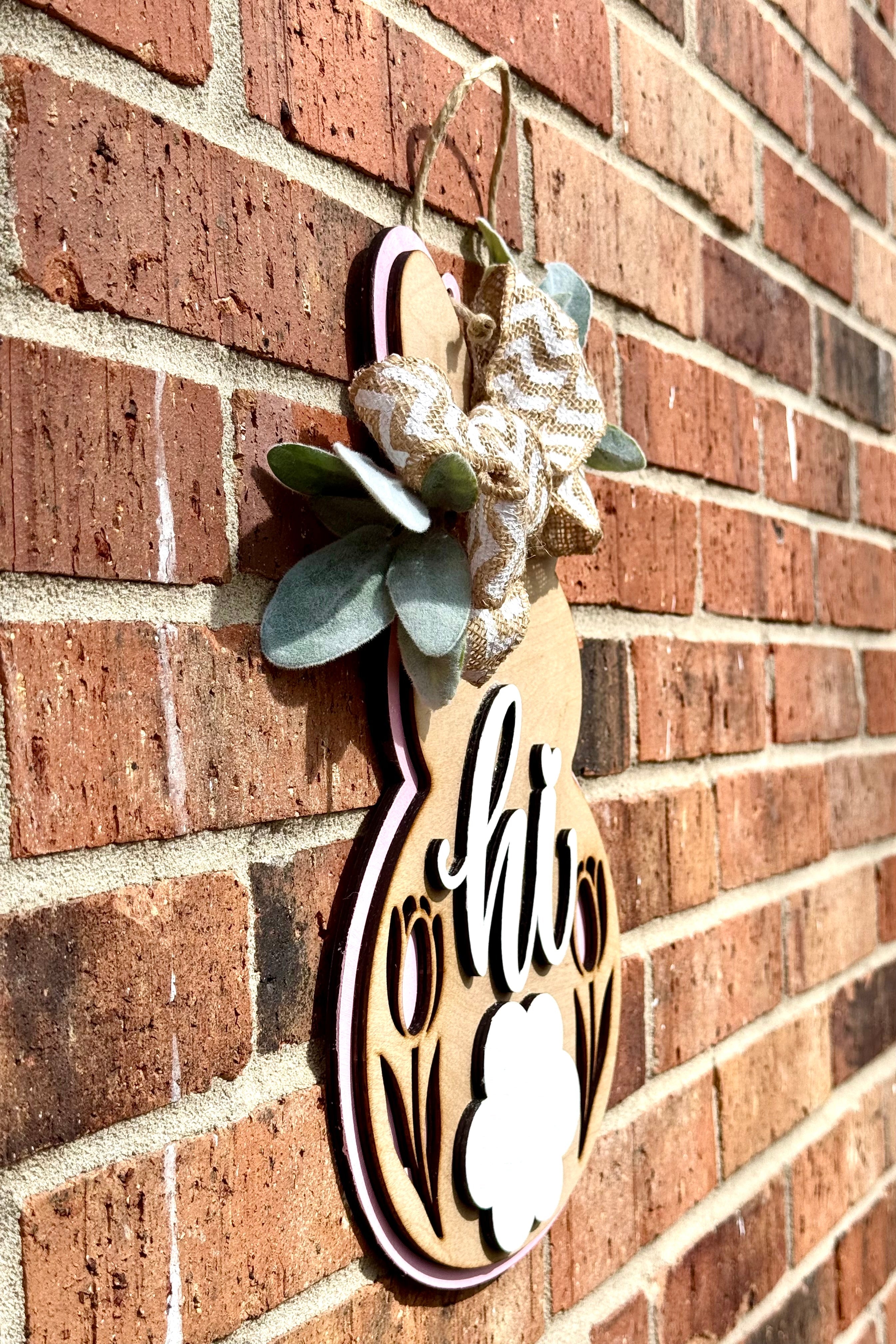 A wooden bunny door hanger with a "Hi" message, bow, and tulip cutouts hangs on a red brick wall. It features farmhouse-style Easter decor.