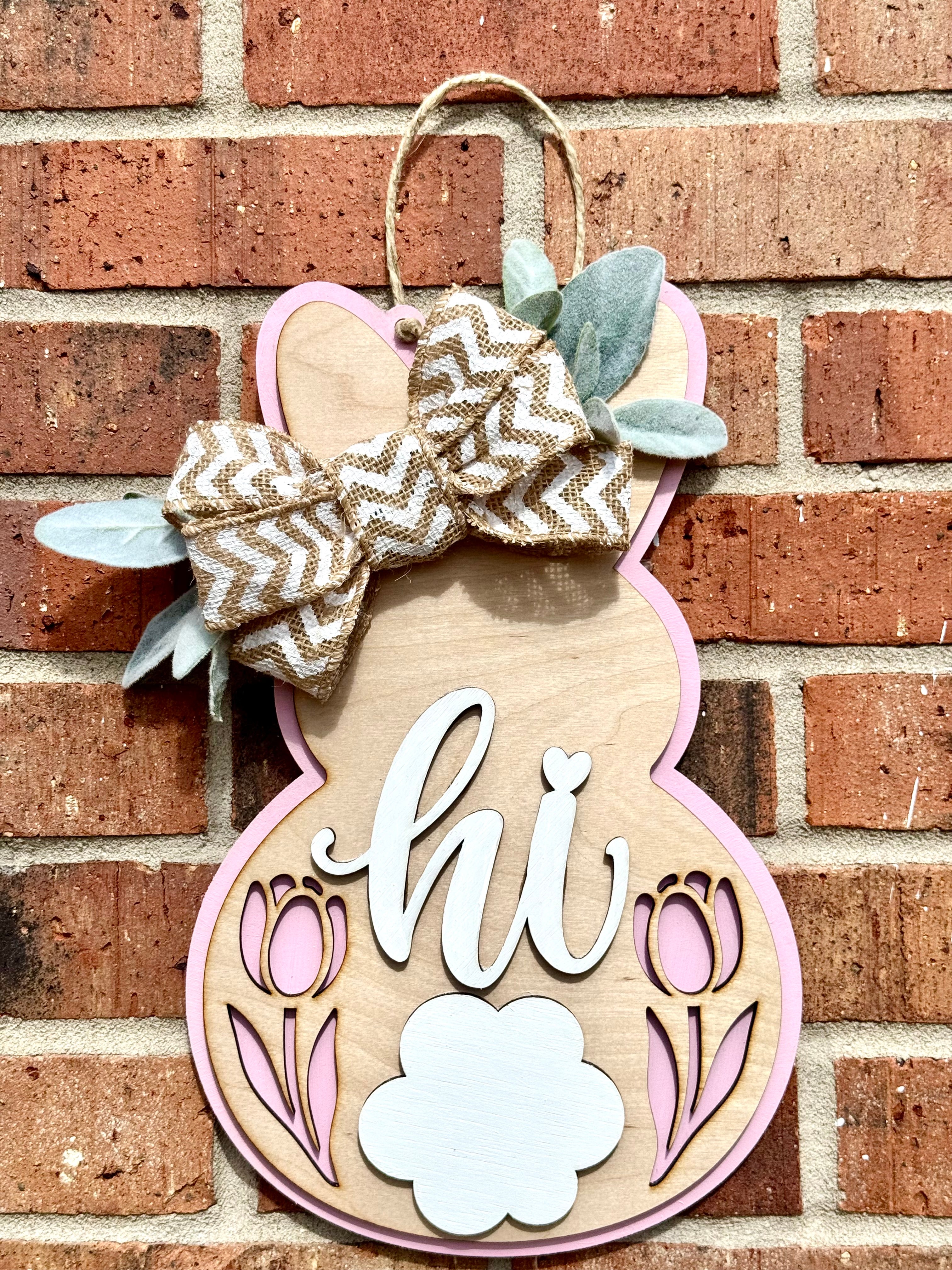 Bunny door hanger with bow, tulips, and 'Hi' sign on brick wall. Wooden Easter decoration with a light pink border and a burlap bow.