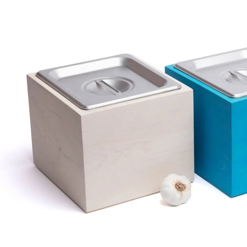 Magnolia wood countertop compost bins with stainless steel interiors, next to a garlic bulb.