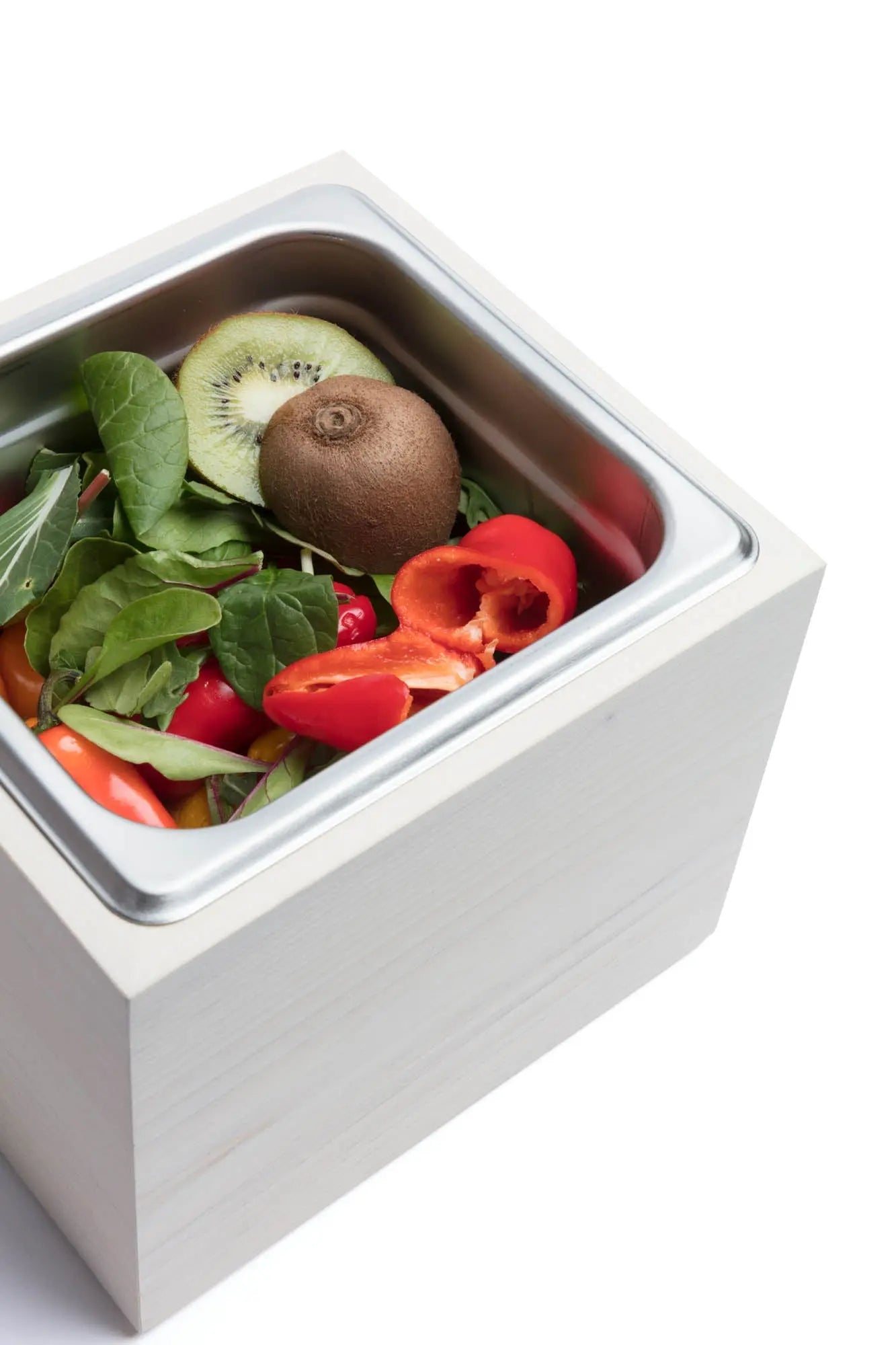 Countertop compost bin with stainless steel insert and magnolia wood exterior filled with kiwi, peppers, and spinach on a white background.