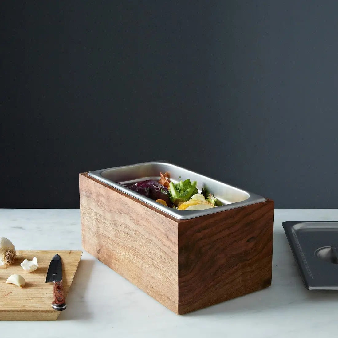Walnut wood countertop compost bin with stainless steel insert, a cutting board, garlic, and a knife on a marble countertop.