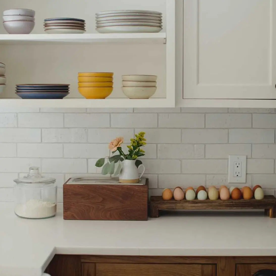 Walnut countertop compost bin with stainless steel interior, kitchen setting with plates, flowers, and eggs.