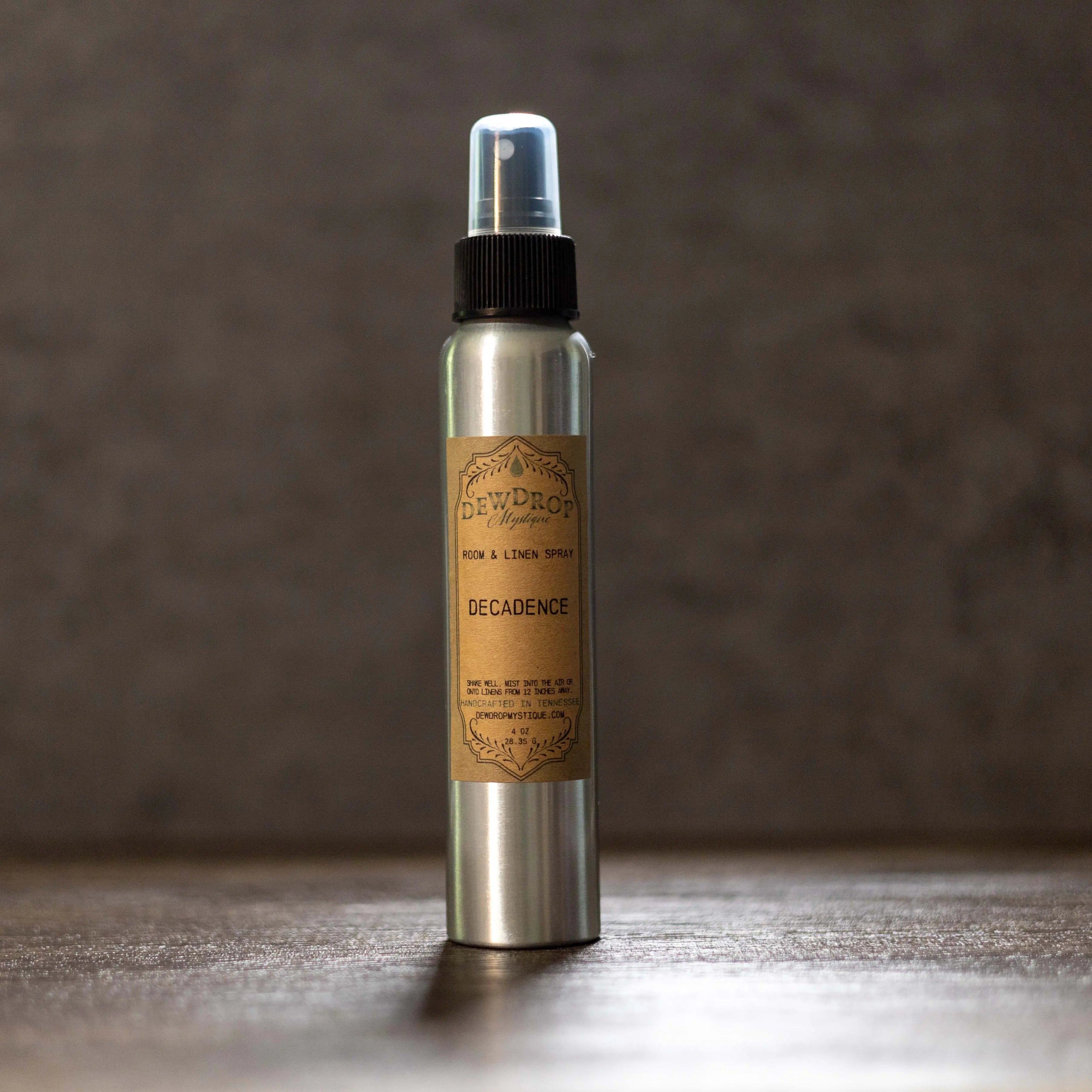Dewdrop Mystique 'Decadence' room spray in a silver bottle with black sprayer and clear cap on a wood surface.