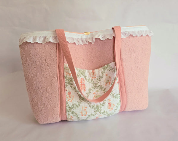 Pink diaper bag with floral pocket, lace trim, and pink straps on a white background. Handmade cottagecore style baby tote.