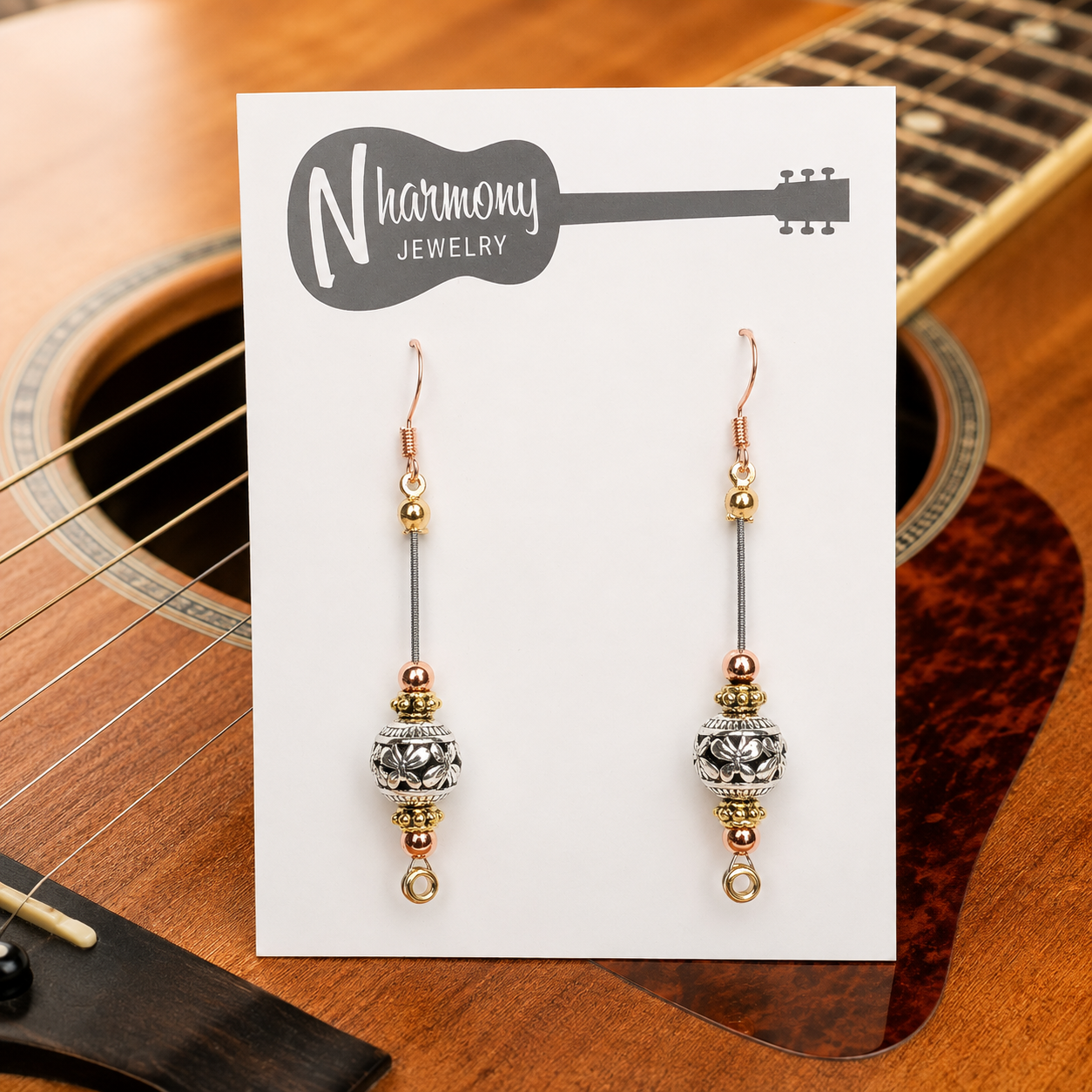Pair of gold and silver beaded earrings on a Harmony Jewelry card with an acoustic guitar in the background.