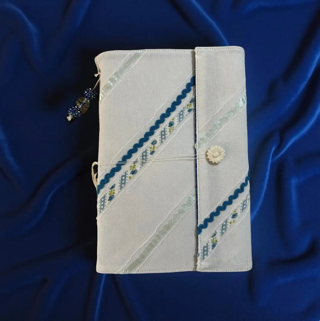 Journal with white fabric cover, ribbon decoration, and snap closure on a blue draped fabric background.