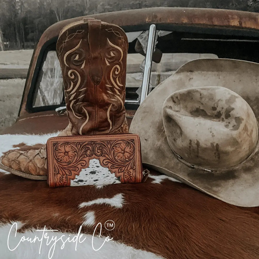 Brown cowboy boots, hat, and tooled leather wallet on cowhide rug in front of a vintage truck.