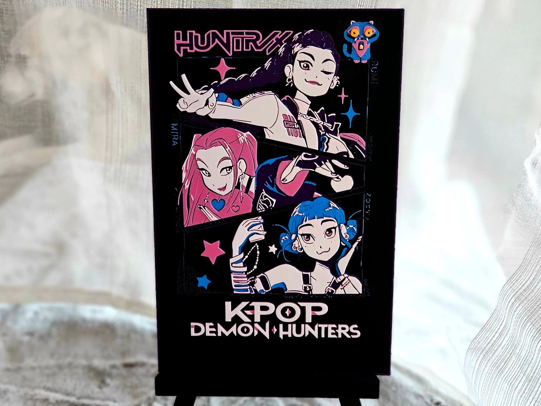 K-POP Demon Hunters poster featuring anime-style illustrations of three characters, a monster and the text: HUNTRX and KPOP DEMON HUNTERS.