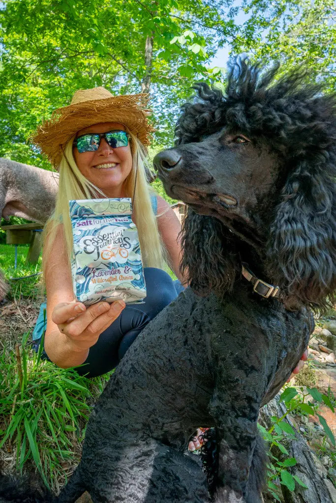 Woman holding lavender and arnica bath salts with a black poodle outdoors