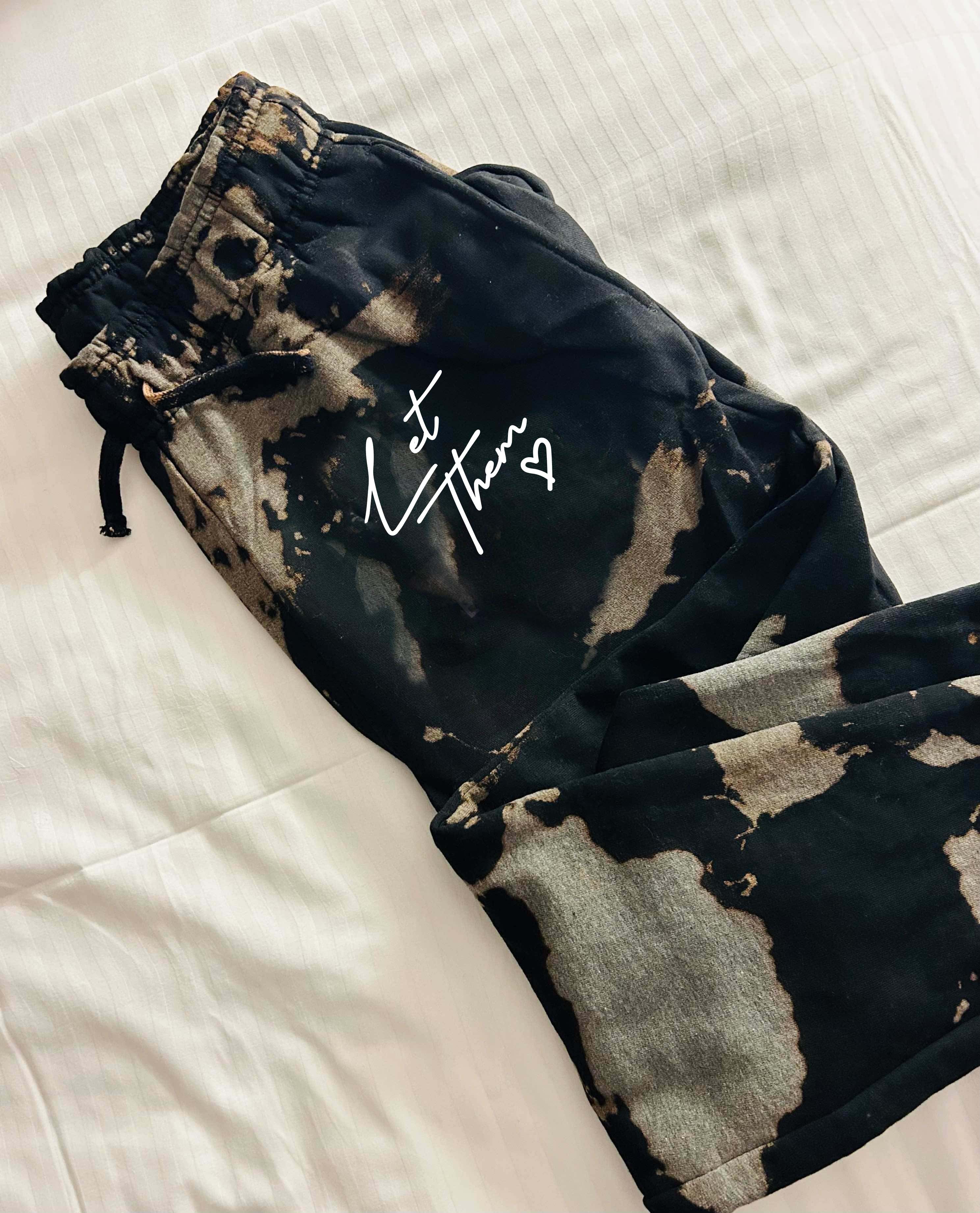 Black and gray tie-dye joggers with drawstring and "Let Them" graphic on a white bedsheet.