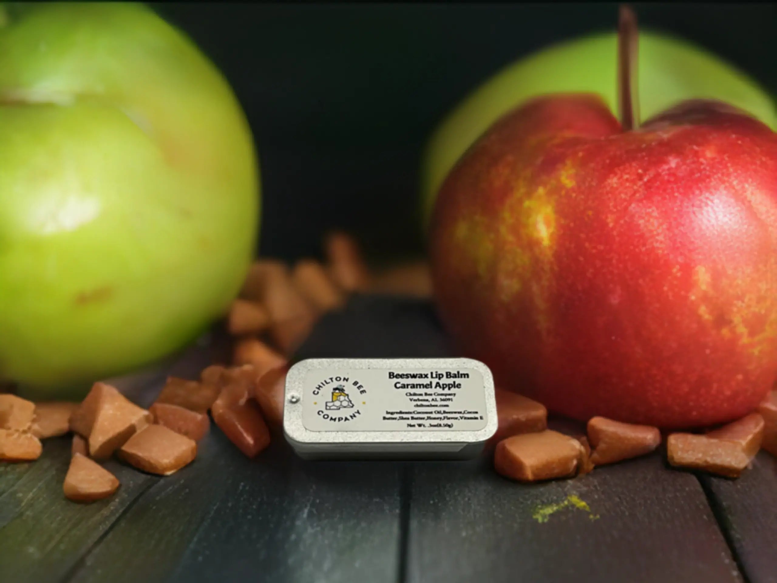 Chilton Bee Caramel Apple lip balm tin with caramel candies and apples on wood surface. Close-up of lip balm.