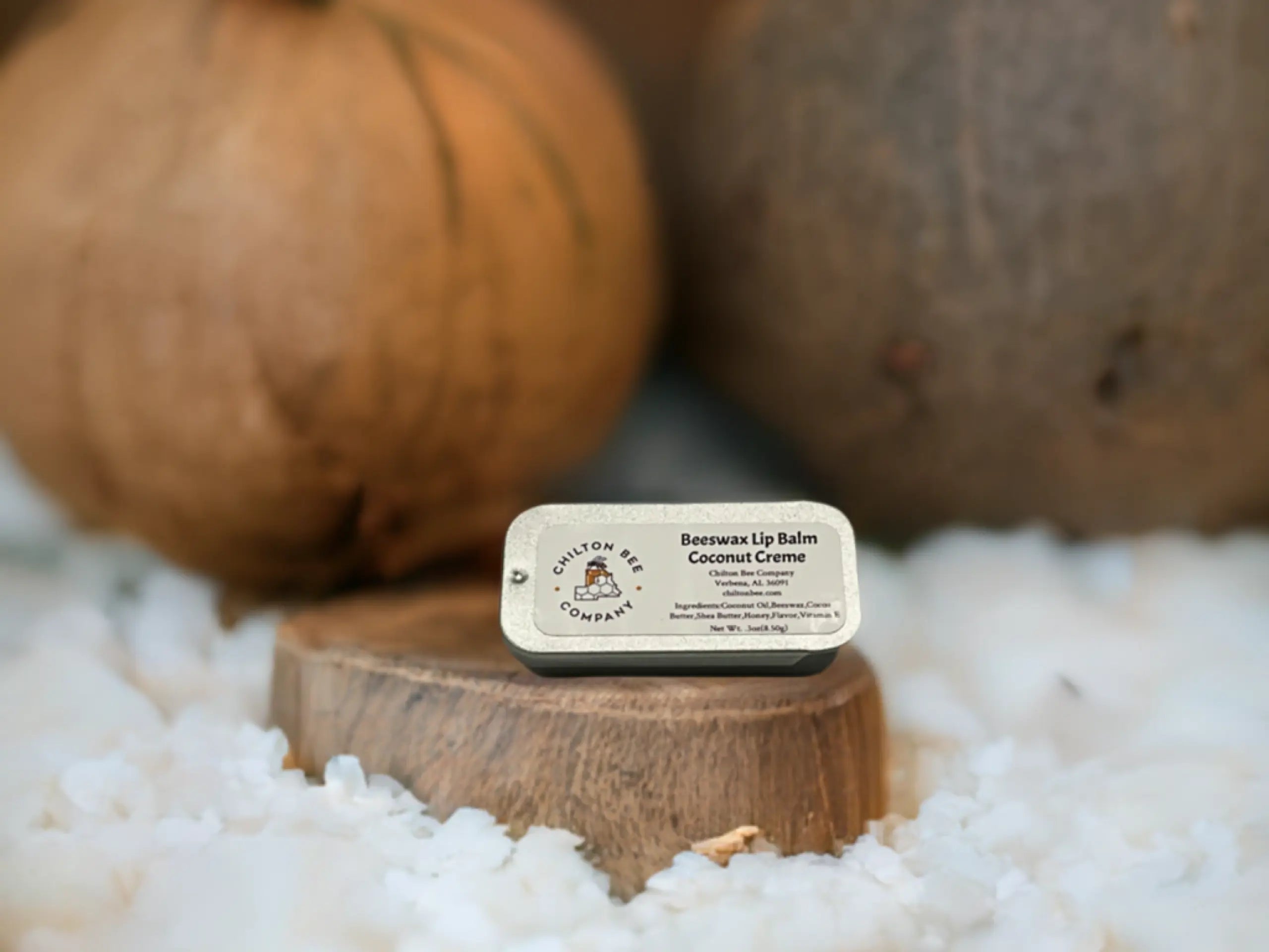 Chilton Bee Company beeswax lip balm in tin with coconut creme label on wood stand, with fuzzy white background