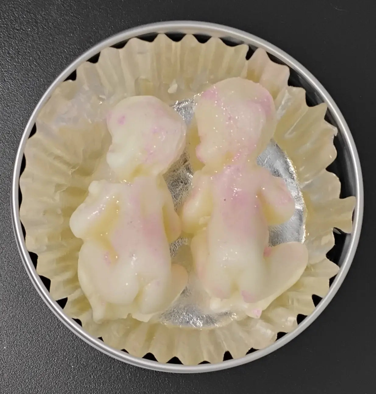 Two baby-shaped lotion bars, white with pink accents, in a paper-lined tin. Handmade lotion bars for moisturizing the skin.