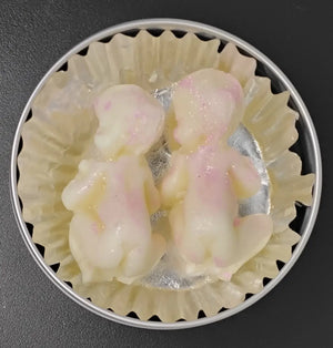 Two baby-shaped lotion bars, white with pink accents, in a paper-lined tin. Handmade lotion bars for moisturizing the skin.