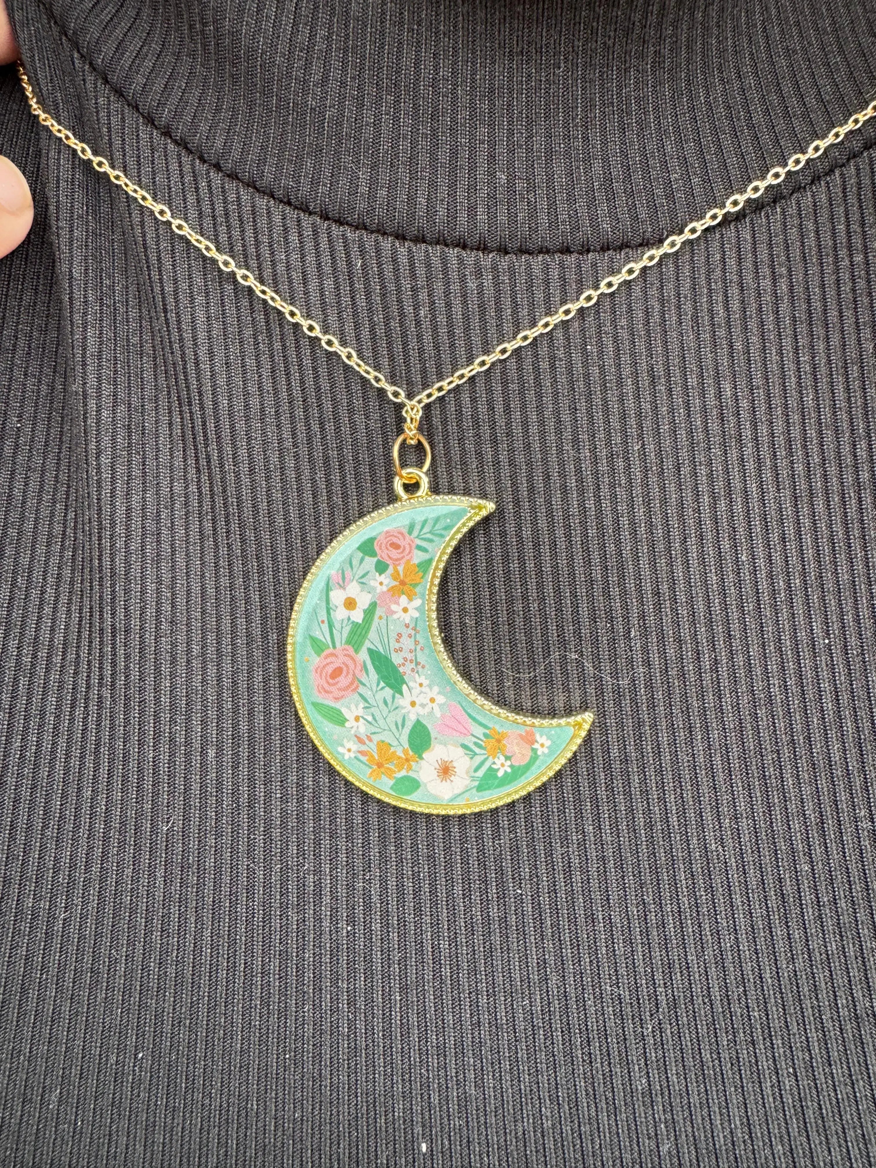 Floral moon pendant necklace on black ribbed fabric. Gold chain and trim, with pastel flower design inside a teal crescent moon shape.