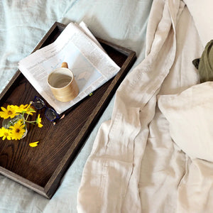 Wooden tray on bed with newspaper, mug, glasses, and flowers. Light blue and off-white bedding visible.