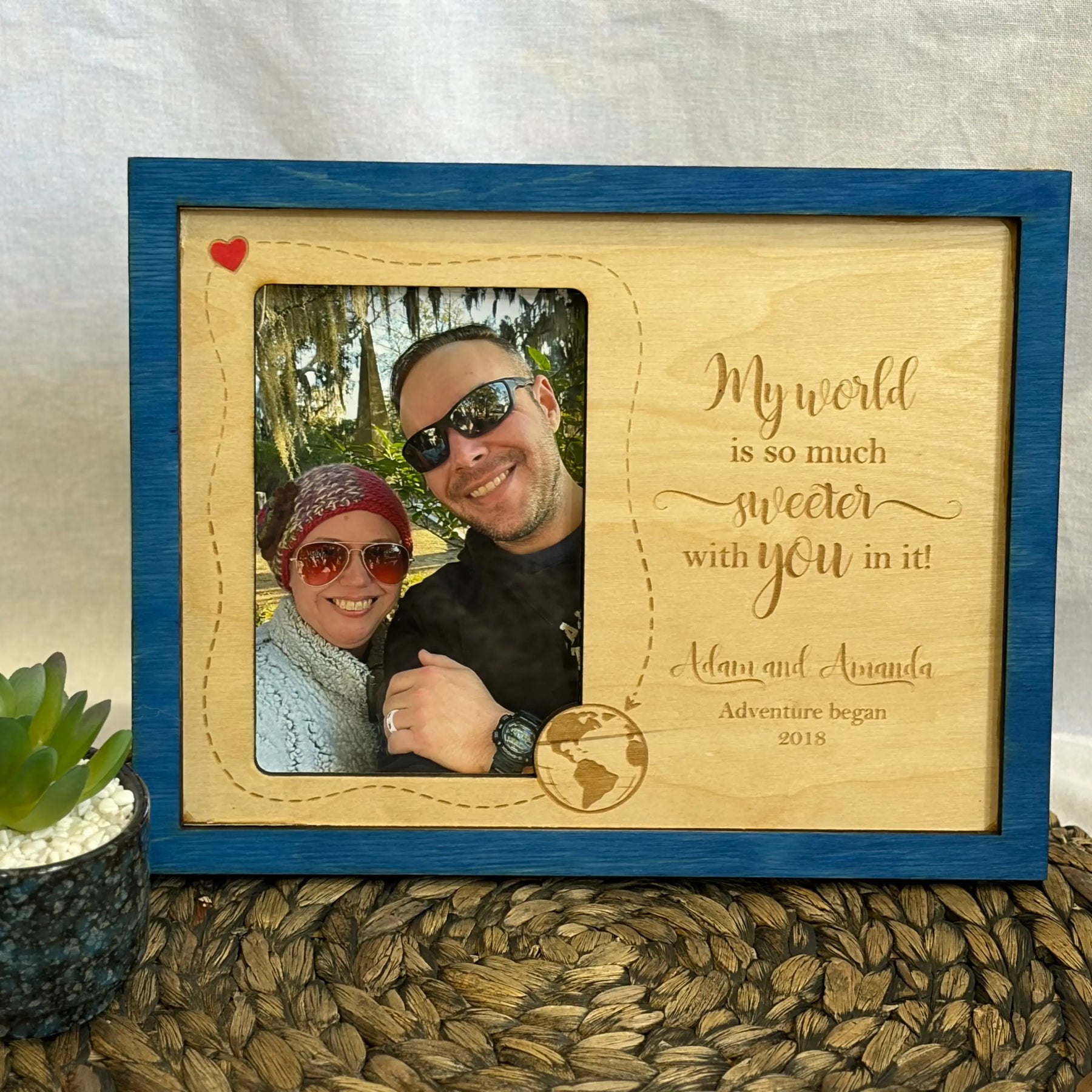 Personalized wooden couple photo frame with engraved love quote, names Adam and Amanda, and date 2018. Blue frame on woven surface.