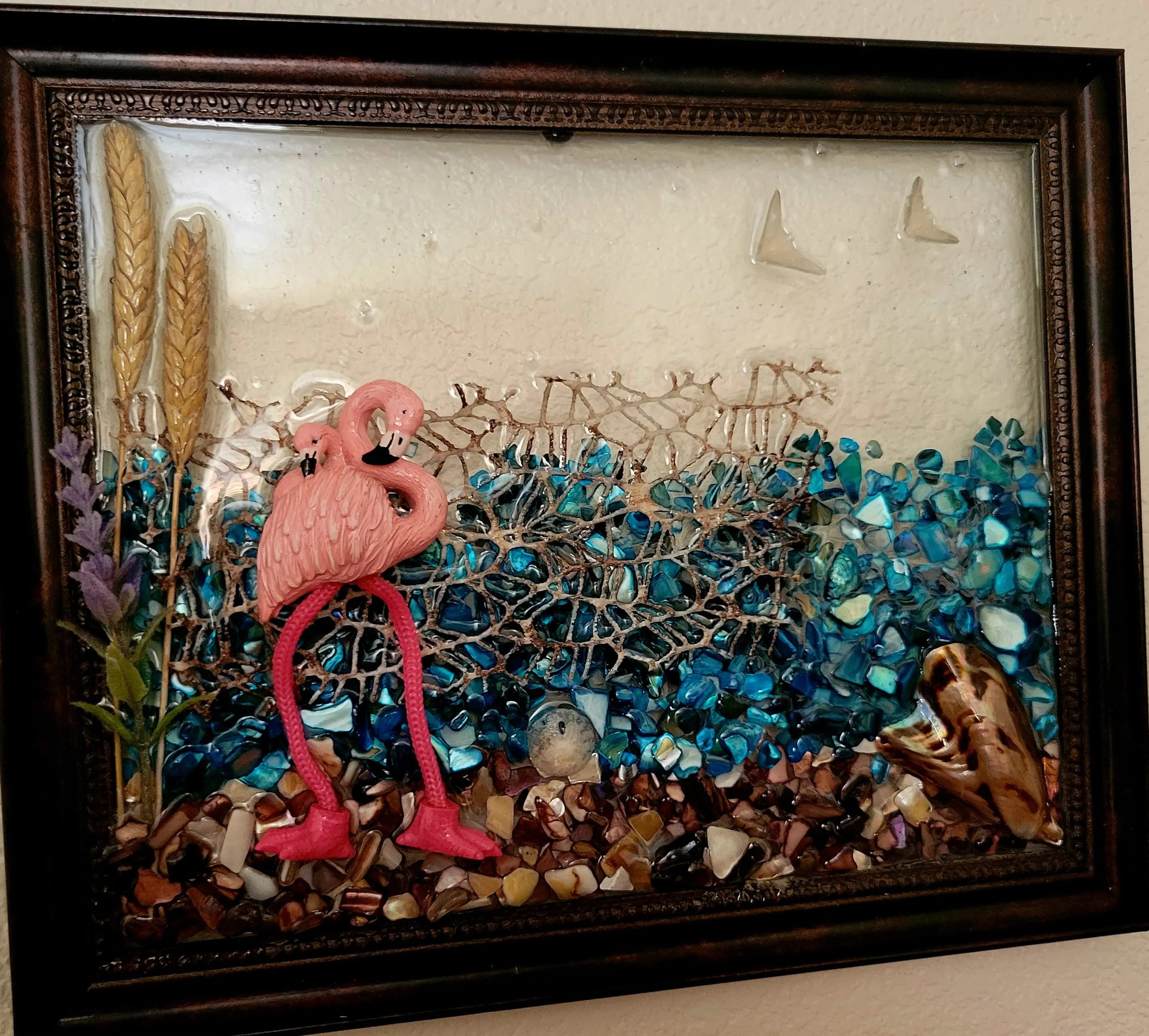 Flamingo coastal wall decor featuring glass resin art with stones and shells in a dark brown frame. 