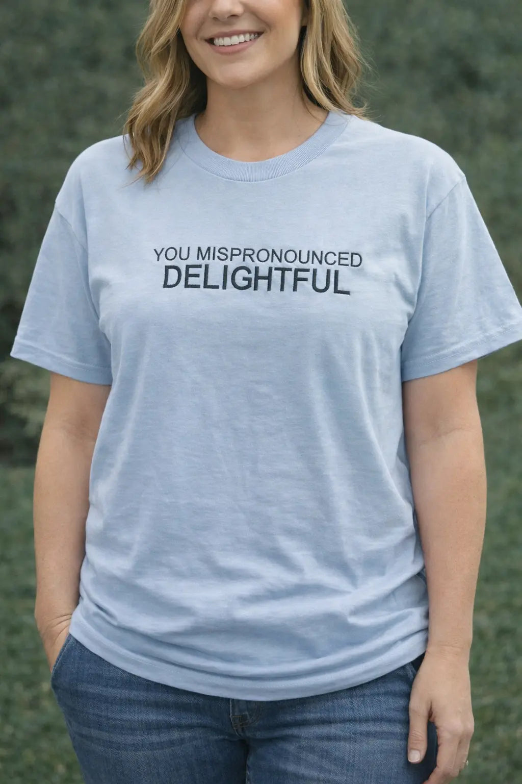 Woman in light blue t-shirt with \"You Mispronounced Delightful\" text, jeans, and green background.