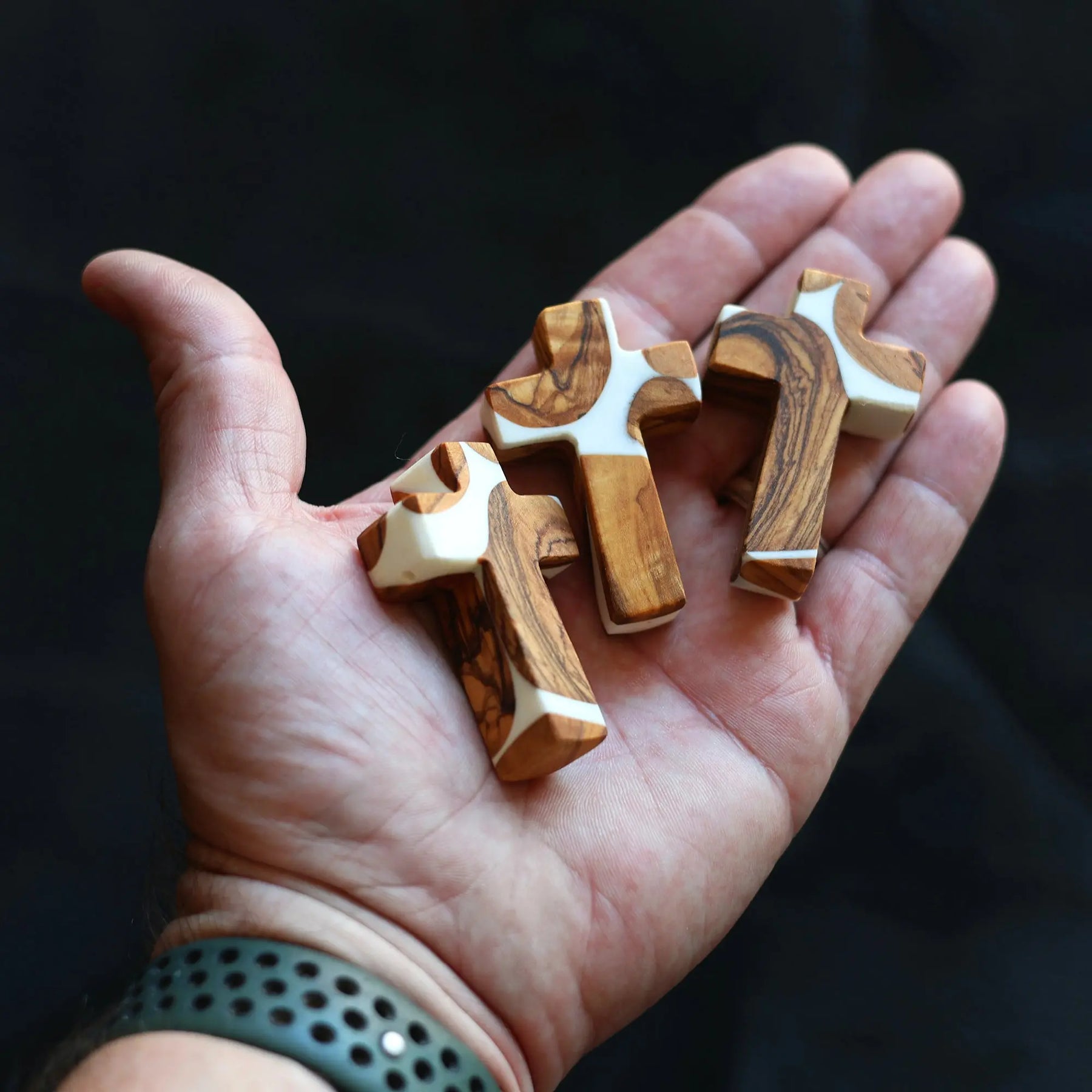 Three olive wood and white resin pocket crosses held in an open hand.