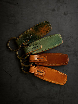 Four leather keychains with antique brass rings in green and brown on a dark background.