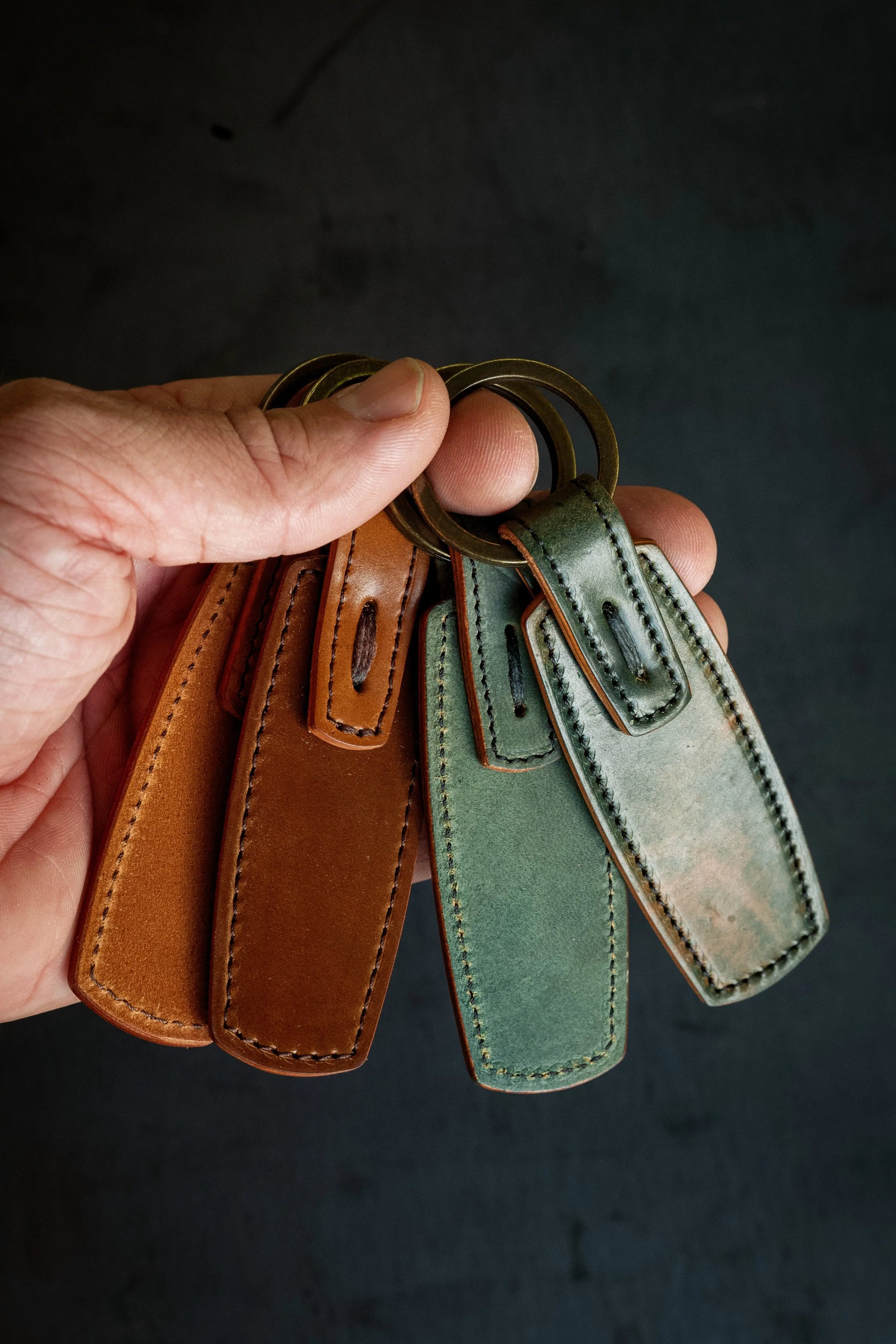 A hand holding a key ring with four leather tags in brown and green, made from Horween Shell Cordovan, attached to antique brass rings.