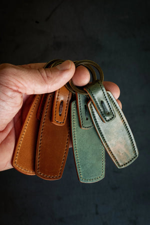 A hand holding a key ring with four leather tags in brown and green, made from Horween Shell Cordovan, attached to antique brass rings.