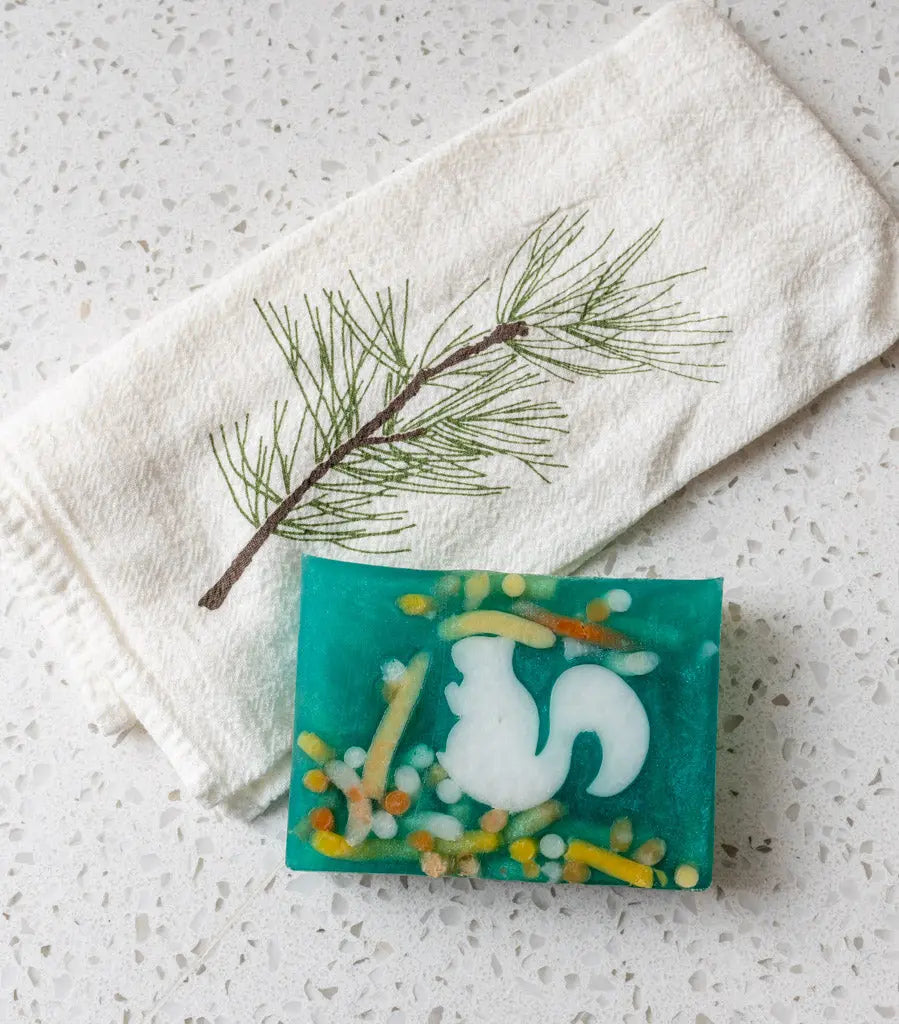 Teal squirrel soap slice with embedded colorful sprinkles and a pine branch embroidered towel.