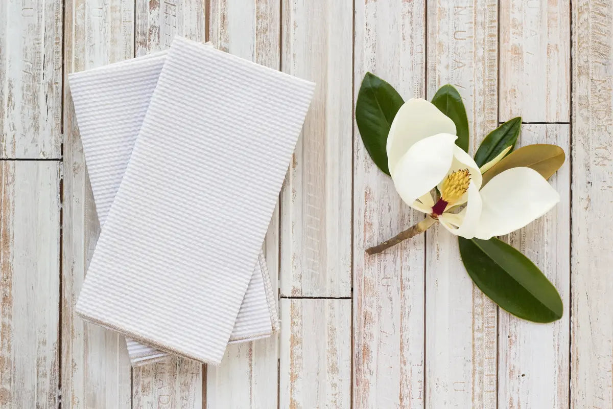 White seersucker dinner napkins folded on a distressed wooden background with a magnolia flower.