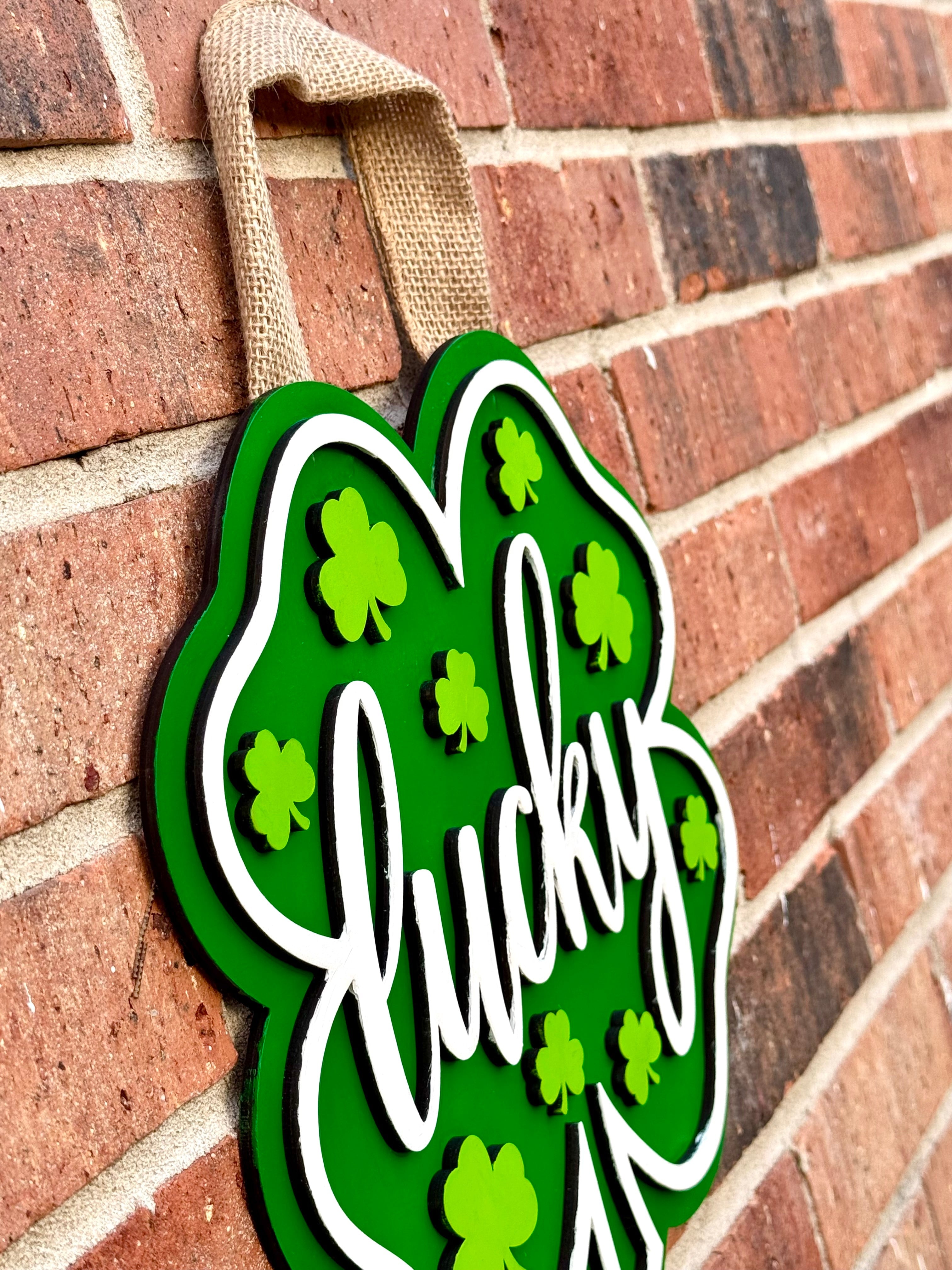 St. Patrick's Day shamrock door hanger: green clover with white "lucky" and burlap hanger on brick wall.