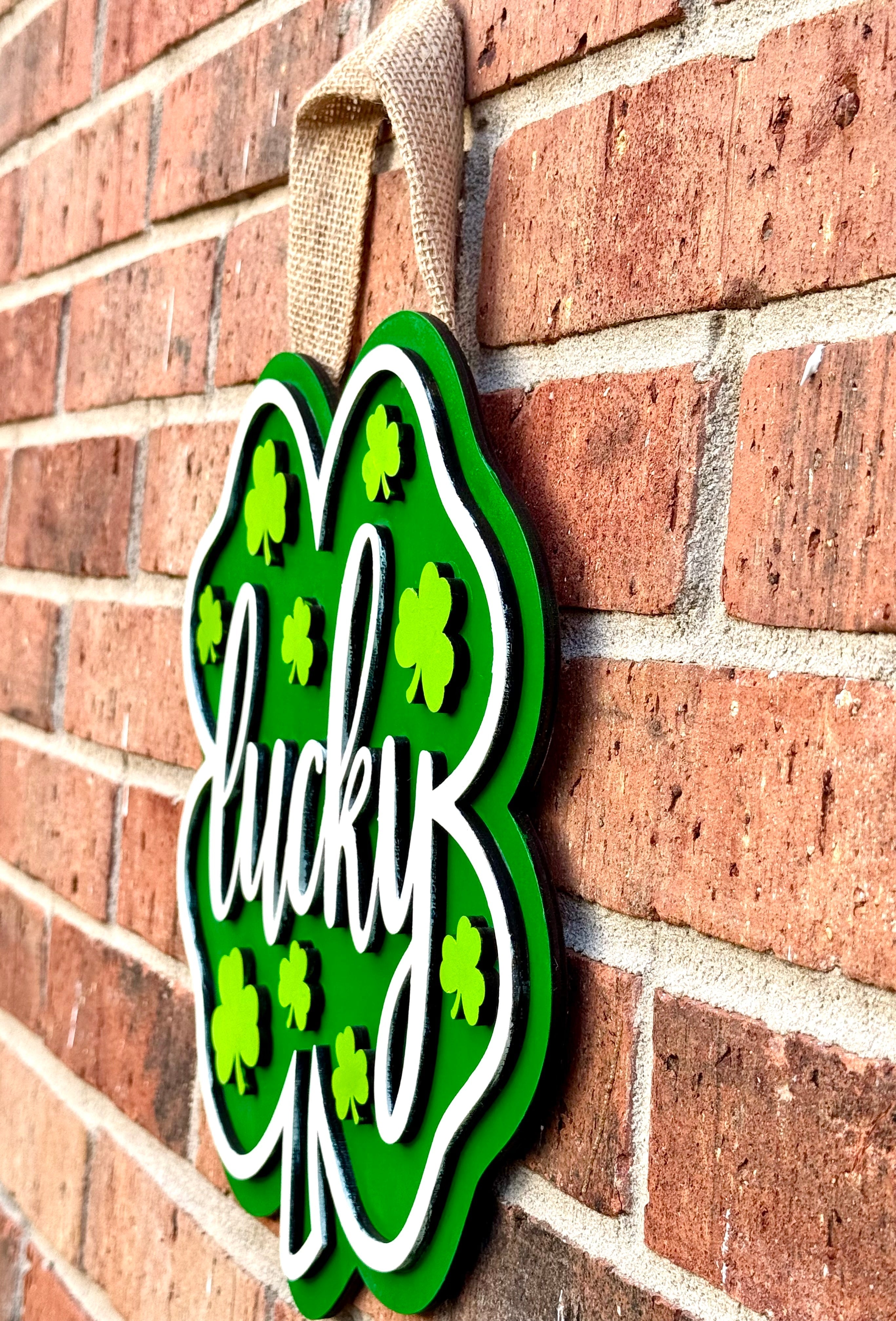 St. Patrick's Day shamrock door hanger with "lucky" text, green cloverleaf design, and burlap ribbon hanging on a brick wall.