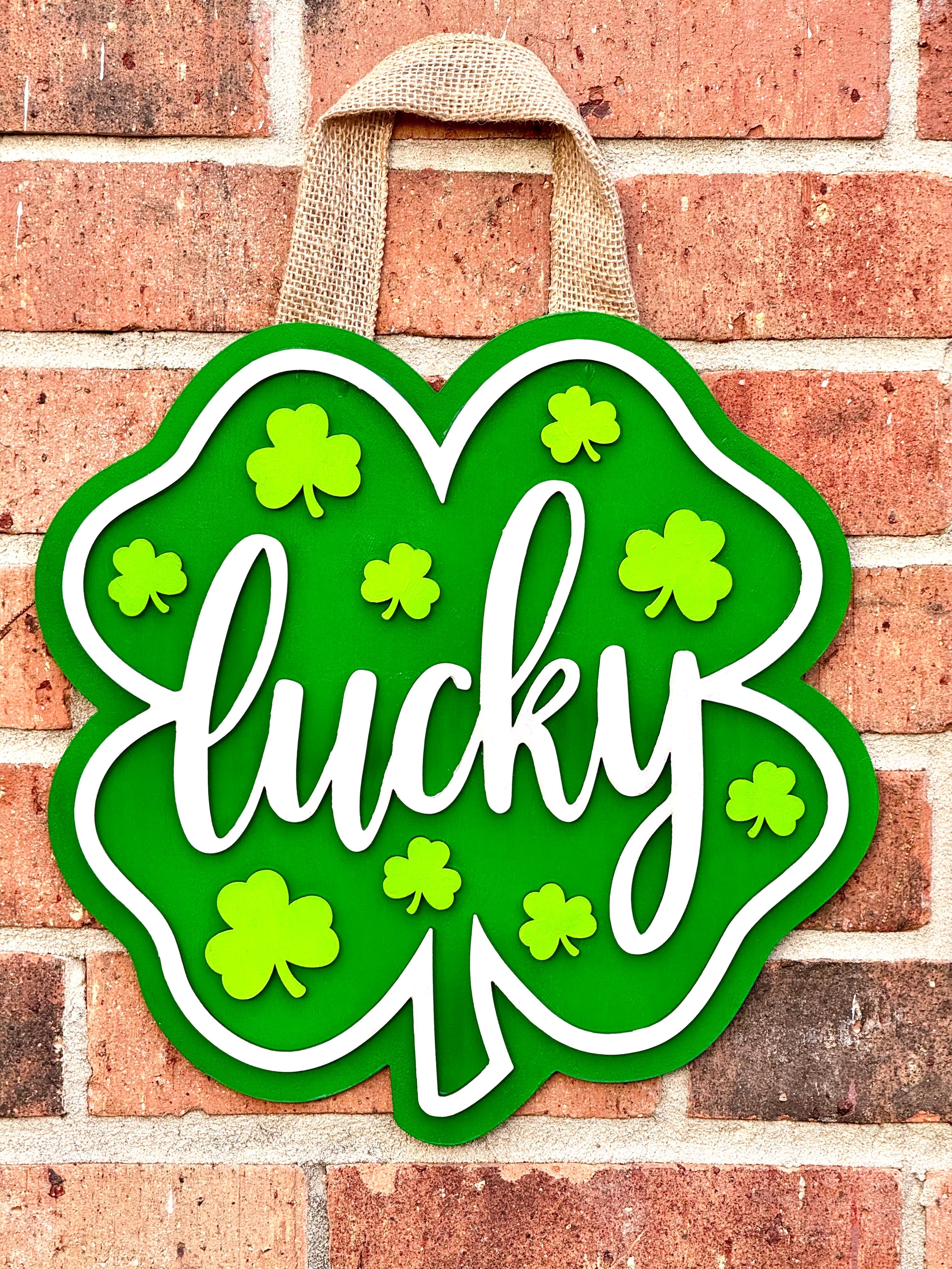Shamrock Door Hanger: Green wood St. Patrick's Day decoration with 'Lucky' text and burlap hanger on brick wall.
