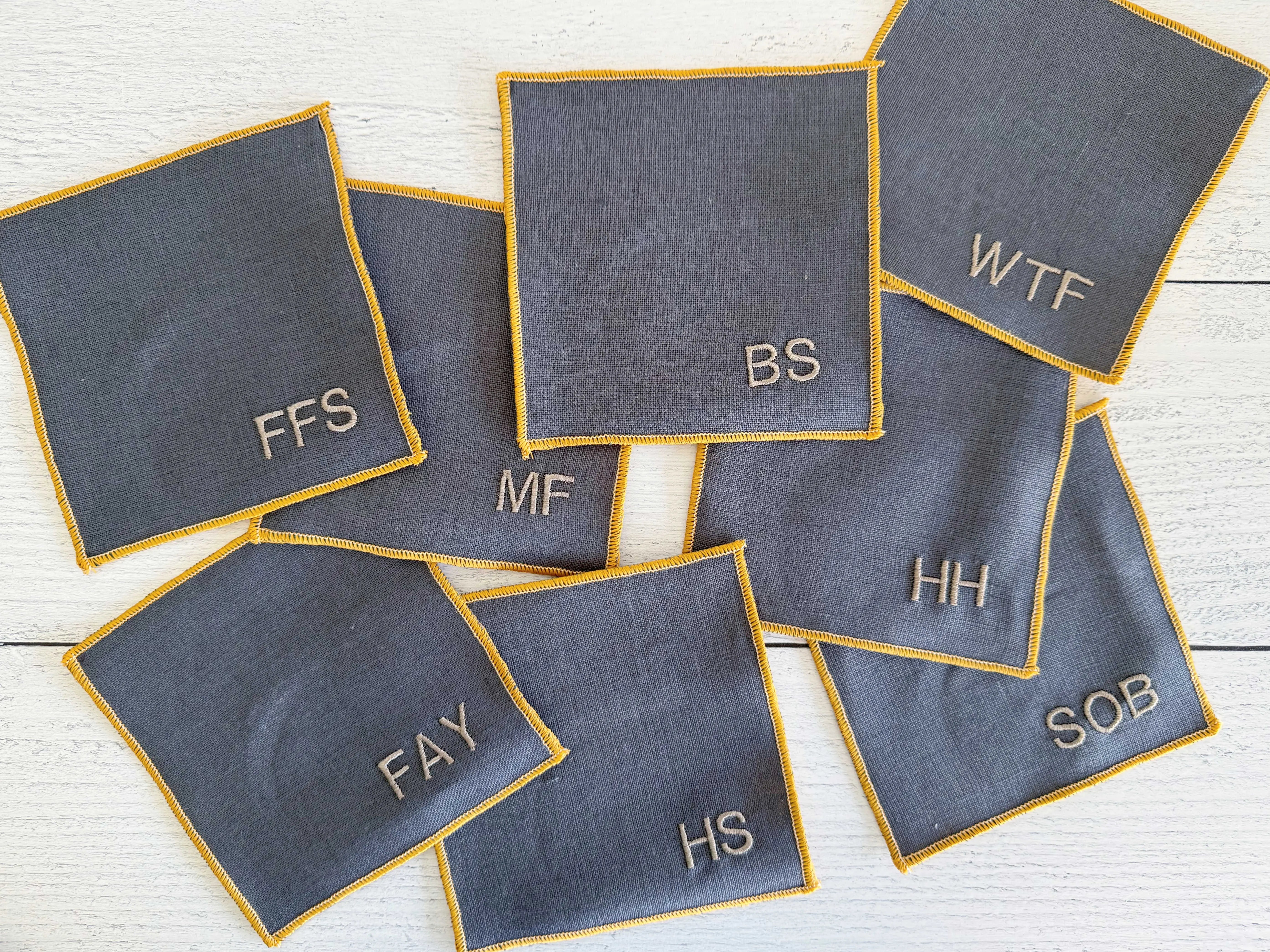 Grey linen coasters with yellow trim and embroidered letters like FFS, WTF, BS, MF, HH, FAY, HS, SOB.